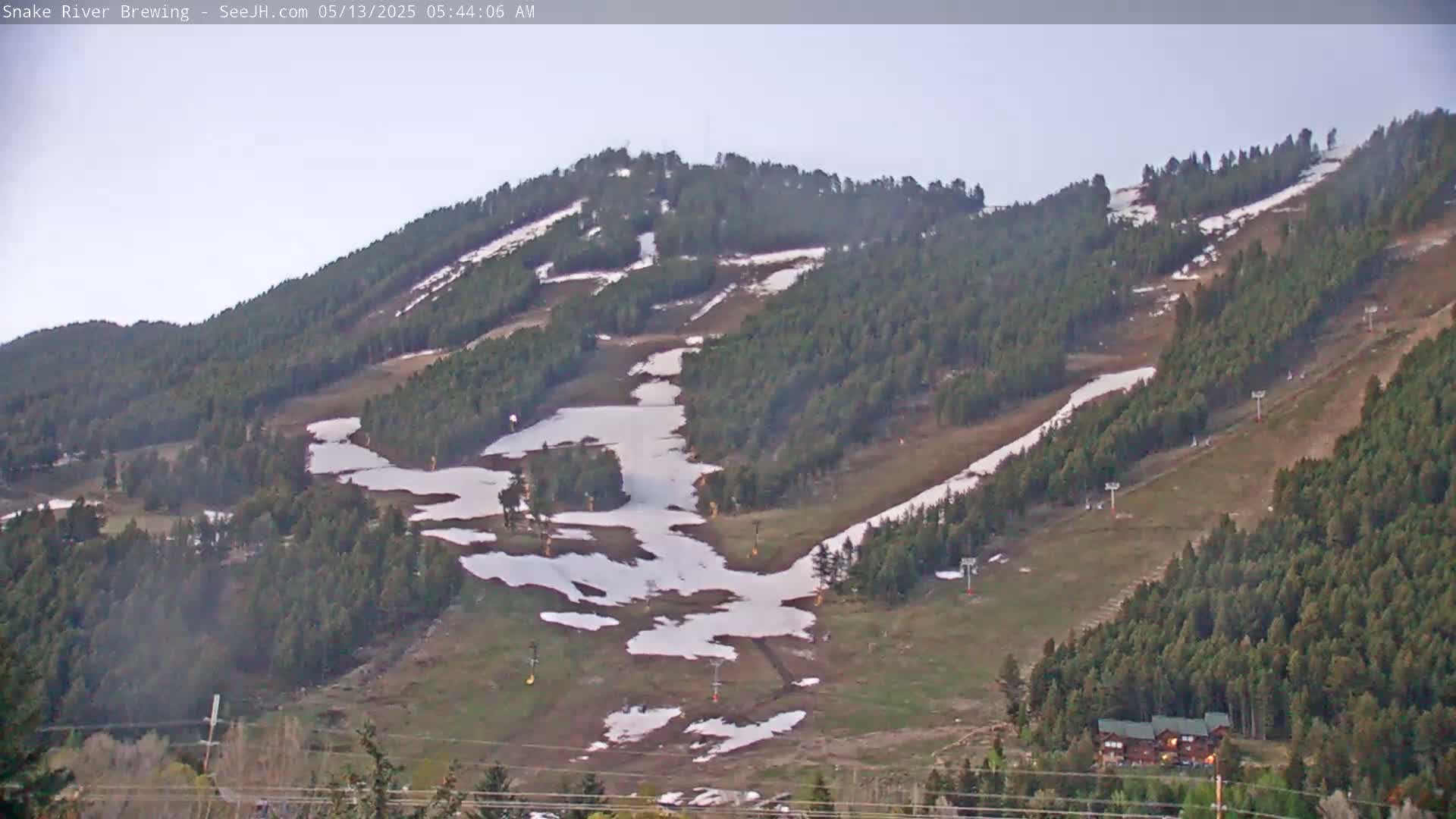 A mostly clear, early morning view of a mountainside with patches of snow, evergreen trees, and ski lifts.