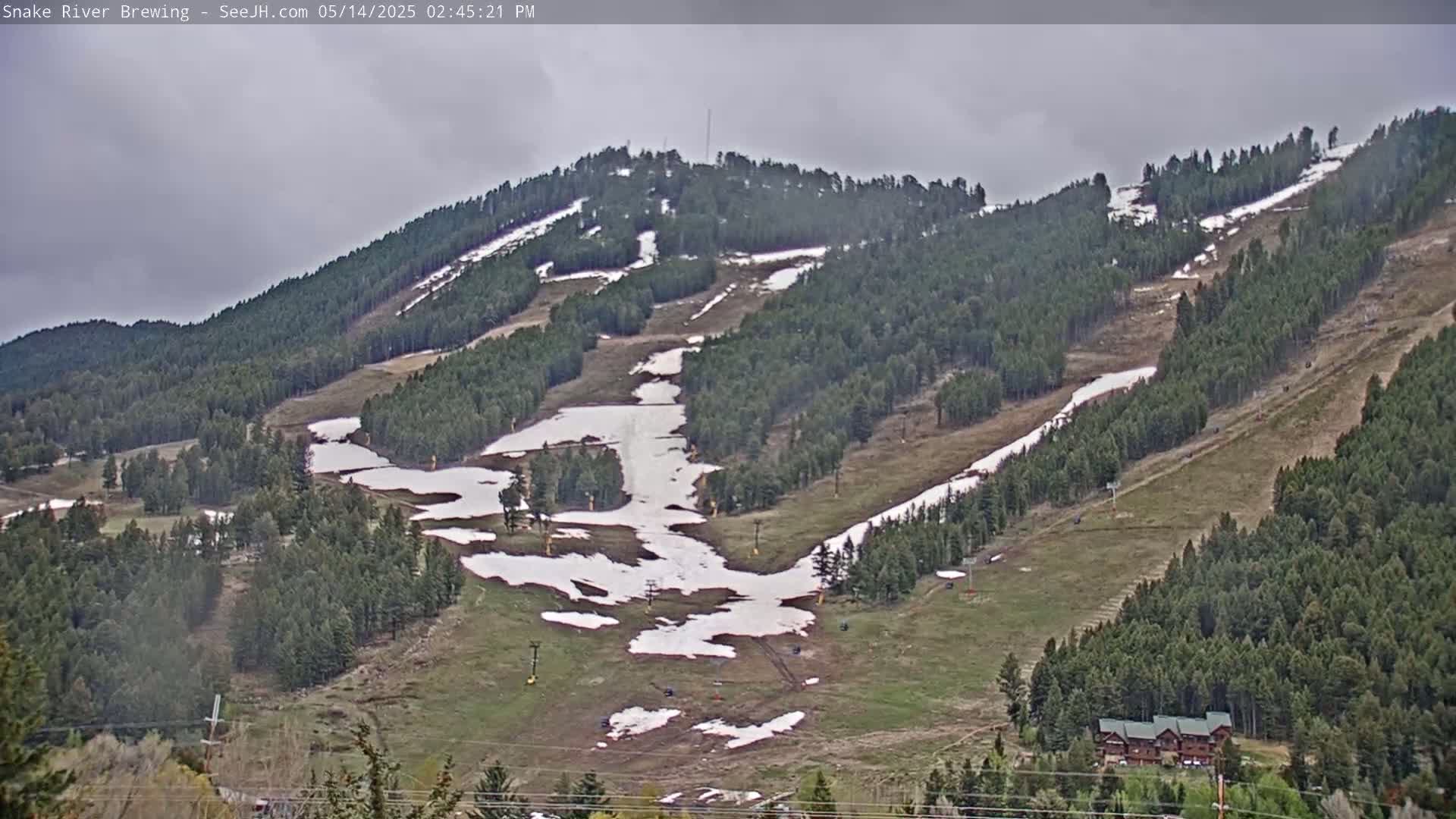A mostly cloudy day reveals a snow-dusted ski slope nestled among evergreen trees, with patches of melting snow visible on the grassy areas.