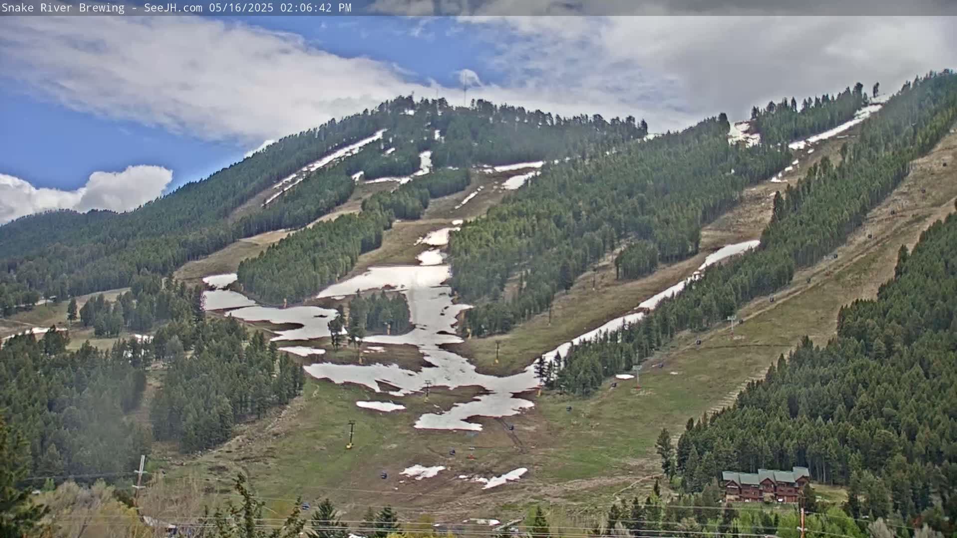 A partly cloudy day reveals a mountainside ski resort with patches of melting snow on the slopes and greening vegetation among the evergreen trees.