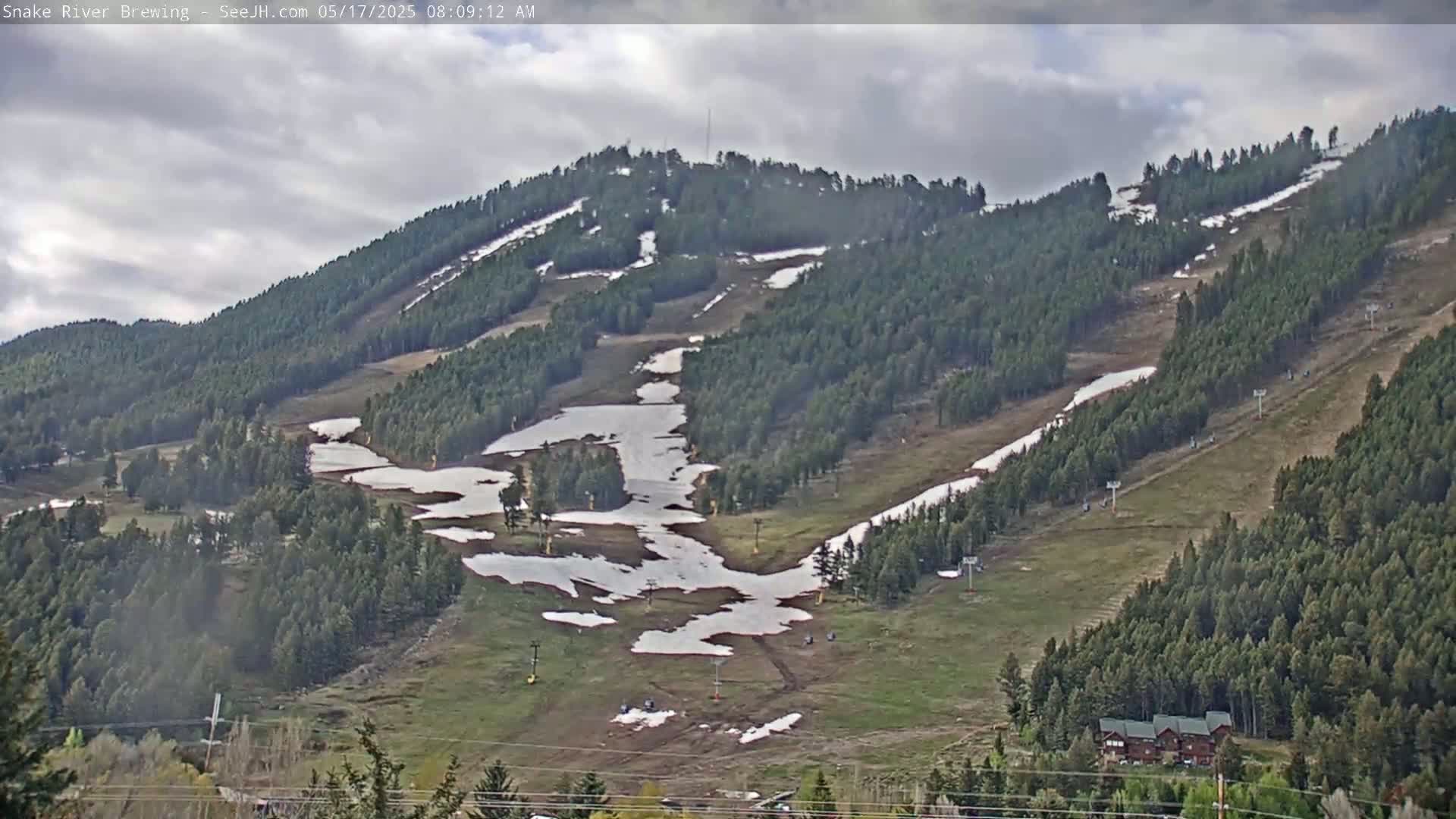 A mostly cloudy day reveals a mountainside ski resort with patches of melting snow on the runs and green grass emerging amongst the evergreen trees.