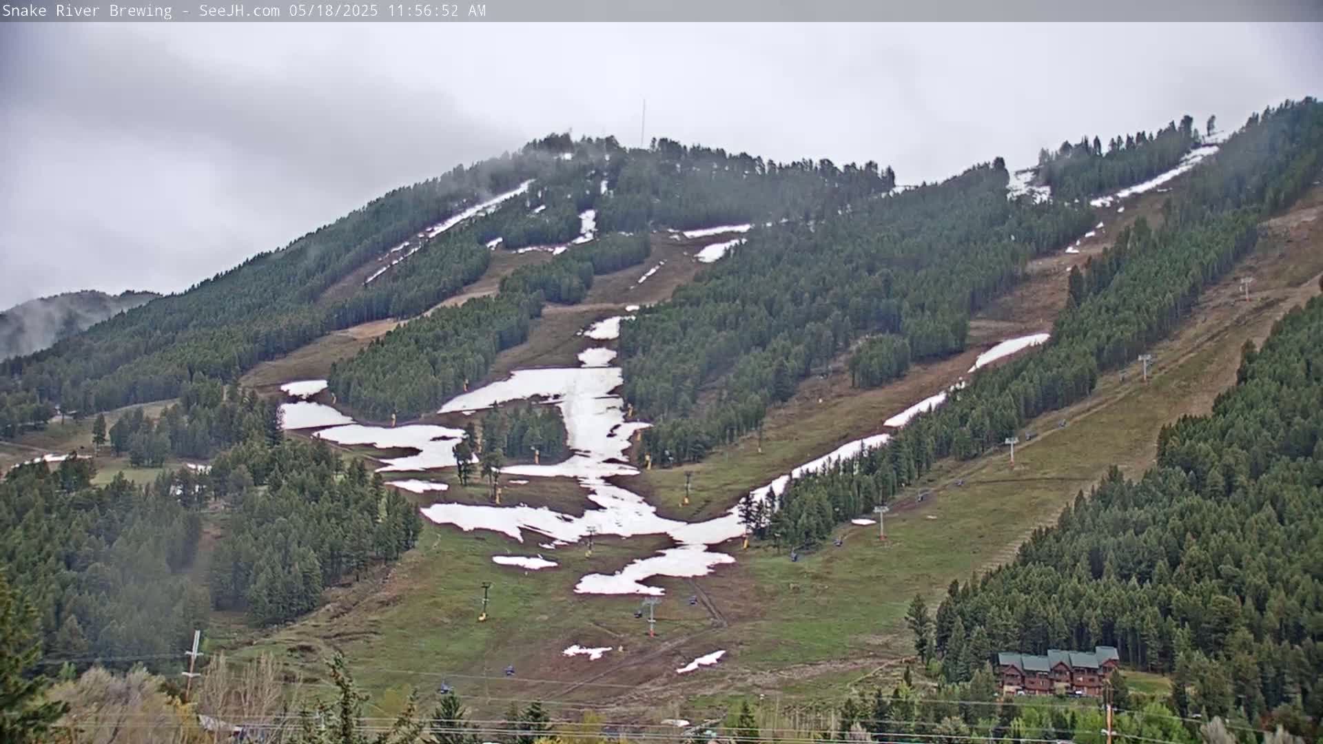A mostly cloudy day reveals a mountainside with patches of snow on the ski slopes, surrounded by evergreen trees and a few buildings at the base.