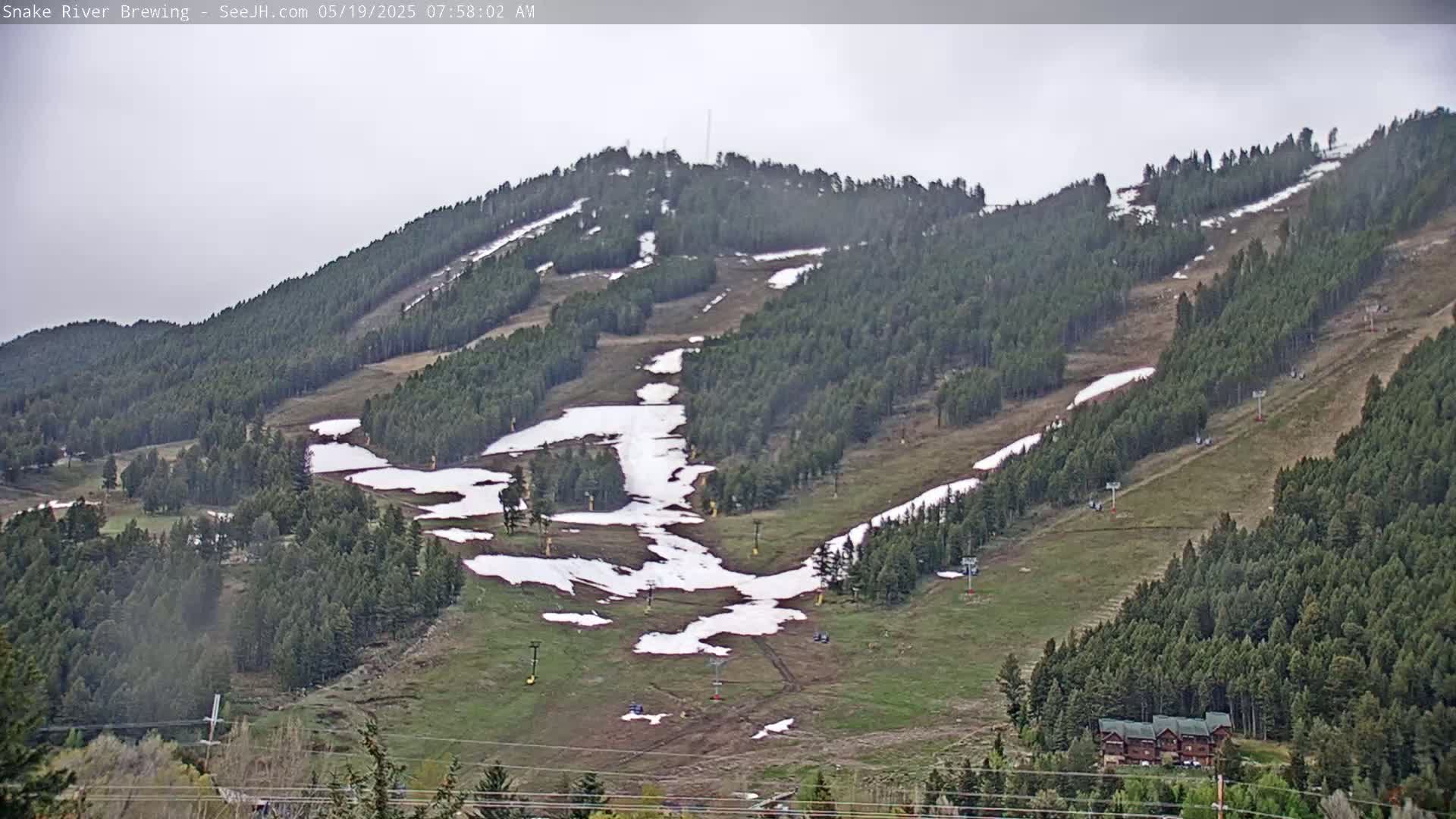 A mostly clear, overcast sky views a ski slope in the spring, with patches of snow remaining on the brown and green grass and trees.