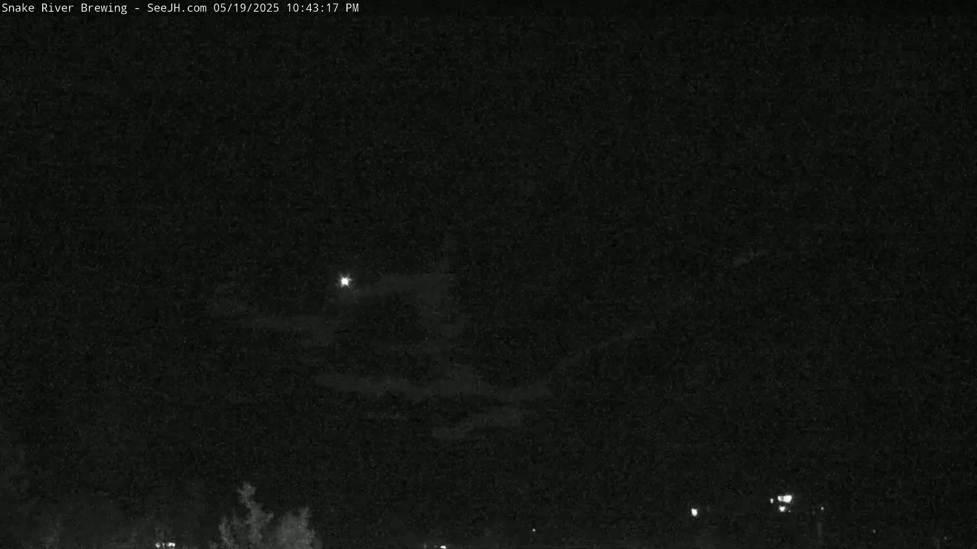 A nighttime view shows a mostly clear sky with a bright light, possibly the moon, and faint clouds, above a dark horizon with a few distant lights.