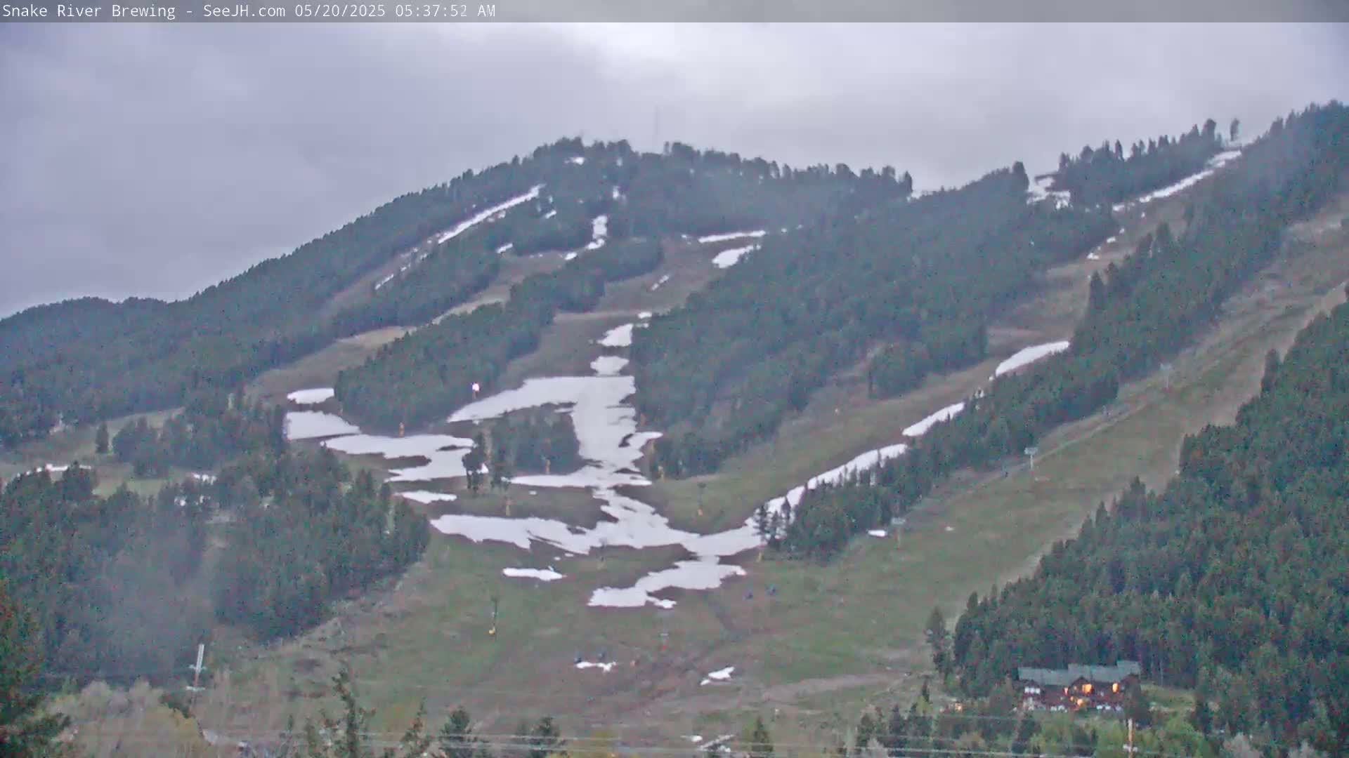 A mostly cloudy day reveals a mountainside with patchy snow on the ski runs and surrounding evergreen trees.