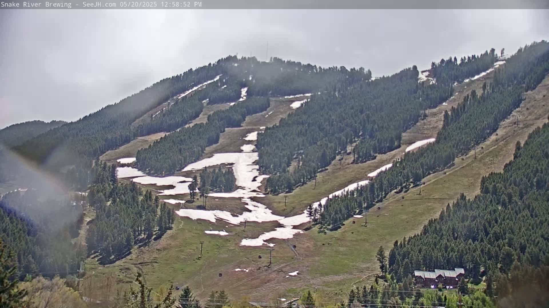 A partly cloudy day reveals a mountainside dotted with evergreens, patches of melting snow, and ski runs.
