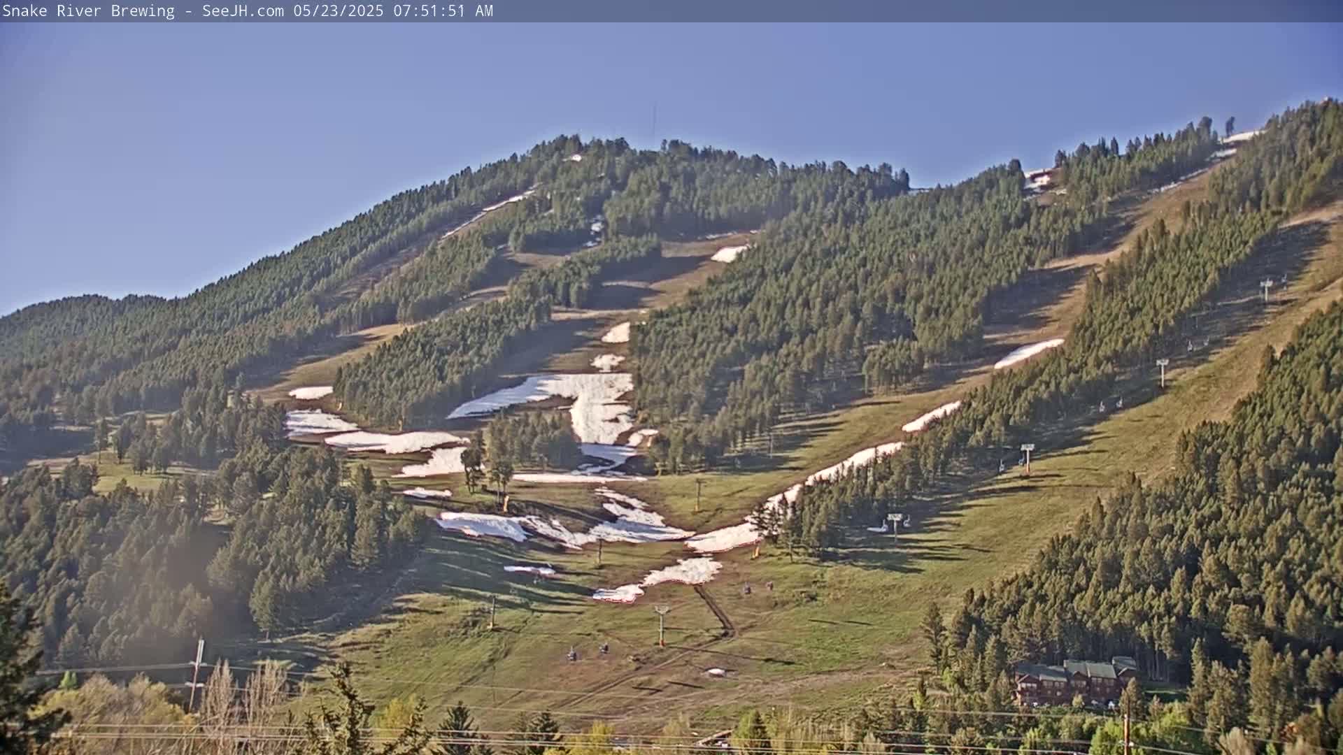 A mostly clear, sunny day reveals a mountainside with patches of remaining snow on ski slopes, surrounded by evergreen trees and grassy areas.