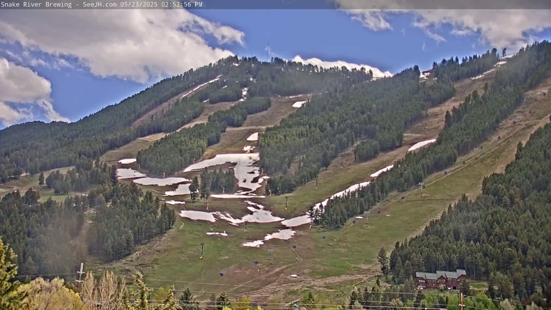 A partly cloudy sky overlooks a mostly green mountainside with patches of snow and several ski lifts.