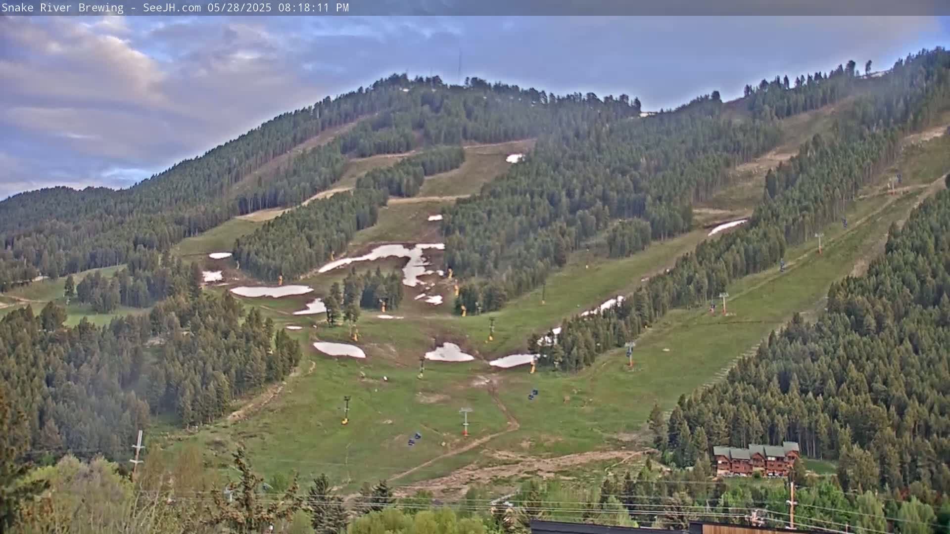 A mostly clear sky overlooks a green mountainside with patches of remaining snow and ski lift infrastructure.