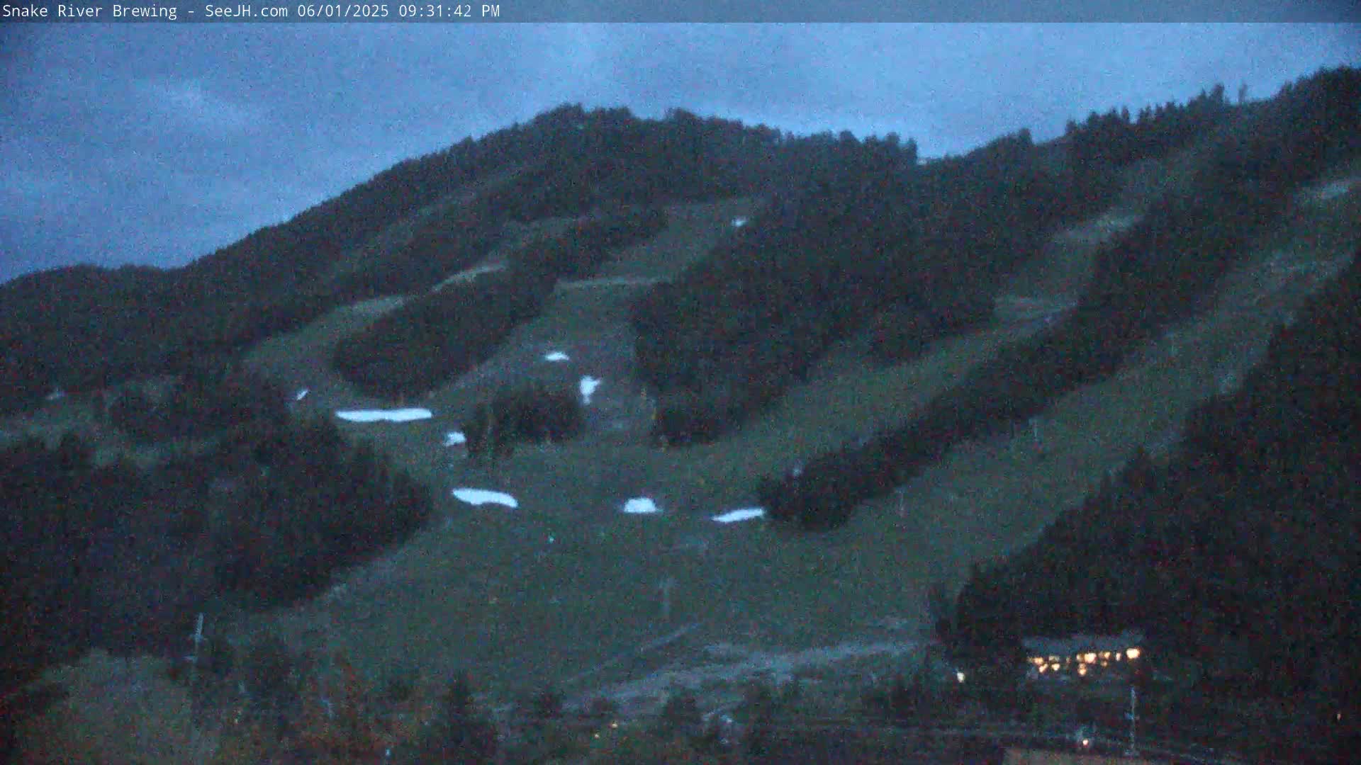 A dark, overcast evening scene shows a hill covered in dark green trees and sparsely scattered patches of snow, with a small building illuminated by lights in the bottom right corner.