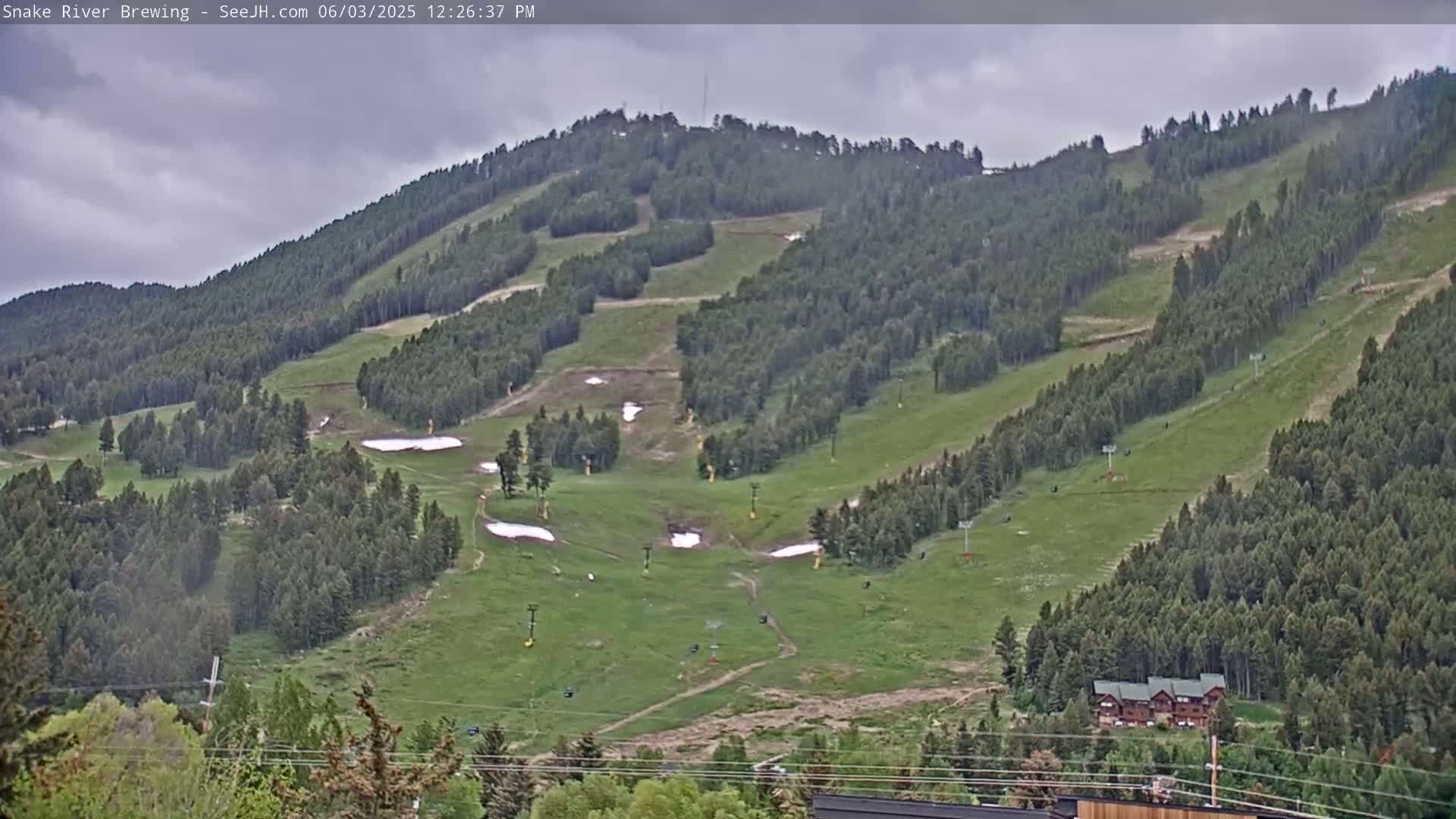 A mostly sunny day reveals a green mountainside with several ski lifts and patches of remaining snow.