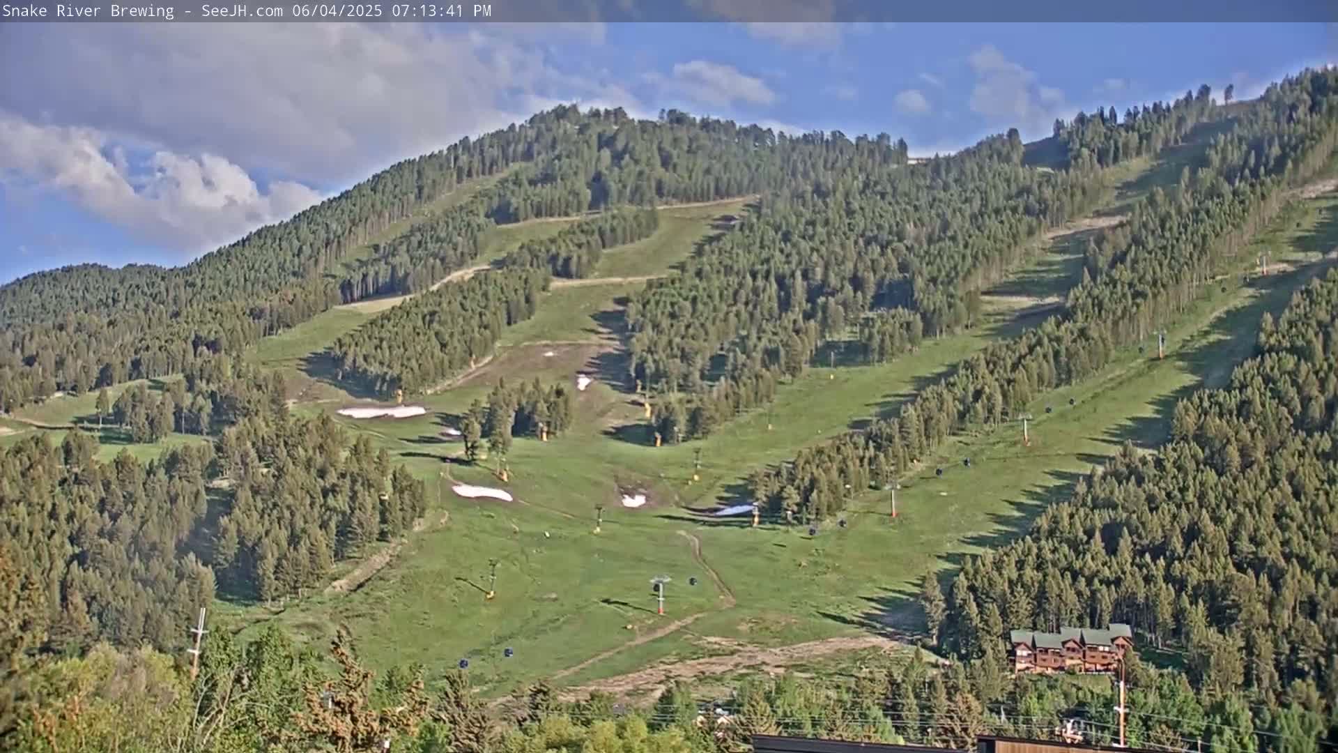 A mostly sunny day reveals a green mountainside with ski lifts and patches of remaining snow.