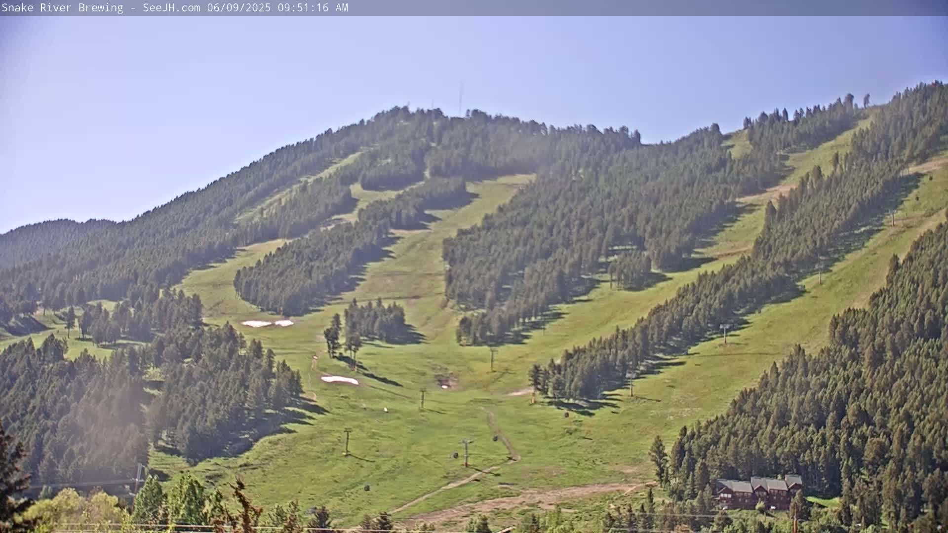 A mostly sunny day reveals a sprawling green hillside dotted with evergreen trees and ski lift infrastructure.