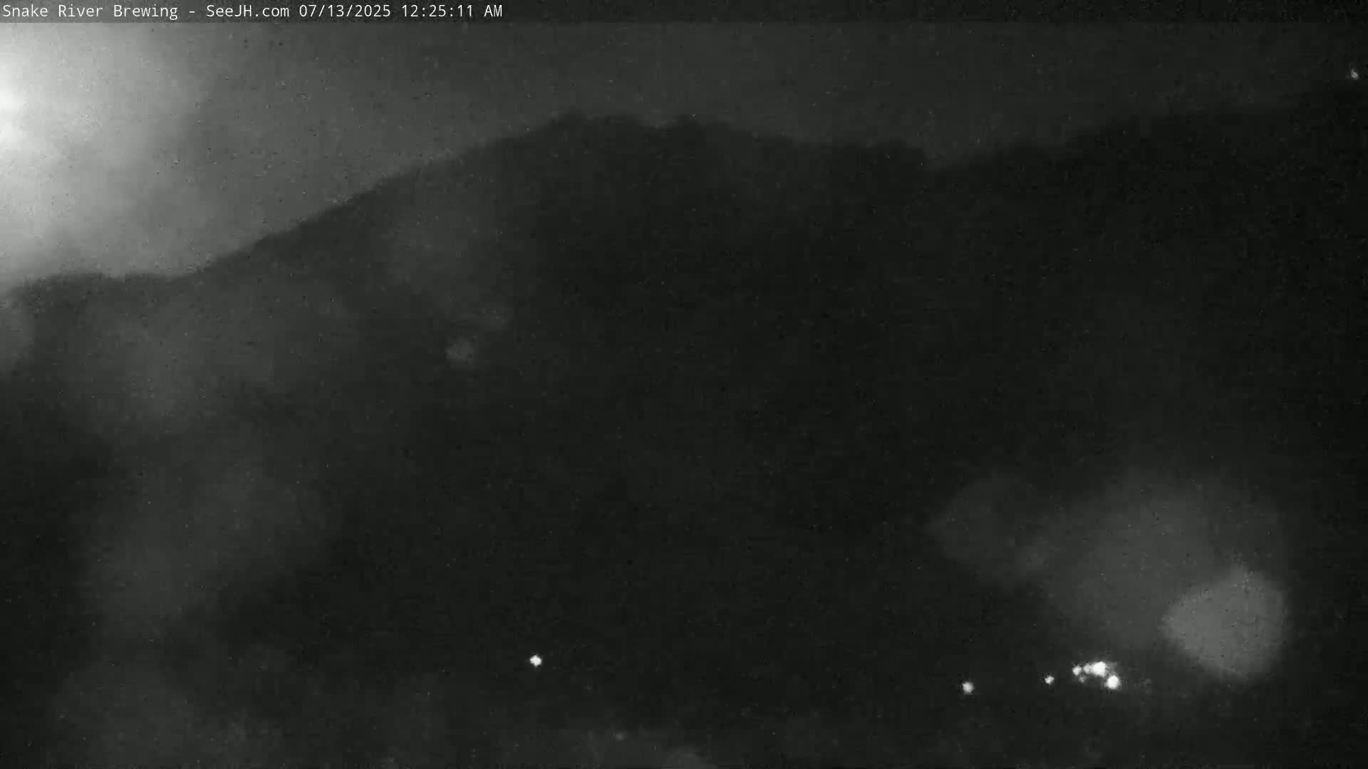 A nighttime, low-light image shows a dark, silhouetted hill or mountain range under a hazy or foggy sky with a few faint lights visible in the lower section of the image.