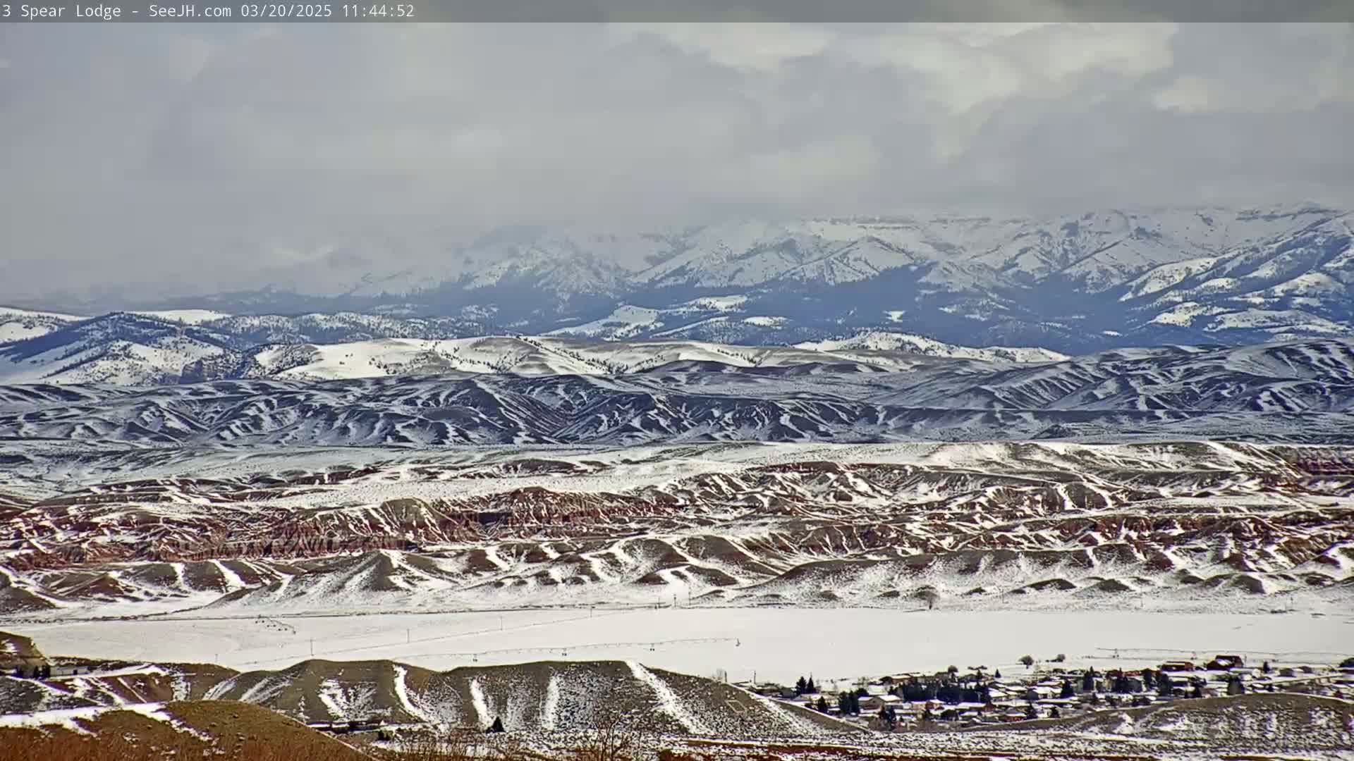 A snow-covered landscape featuring rolling hills, a small town, and a distant mountain range under an overcast sky.