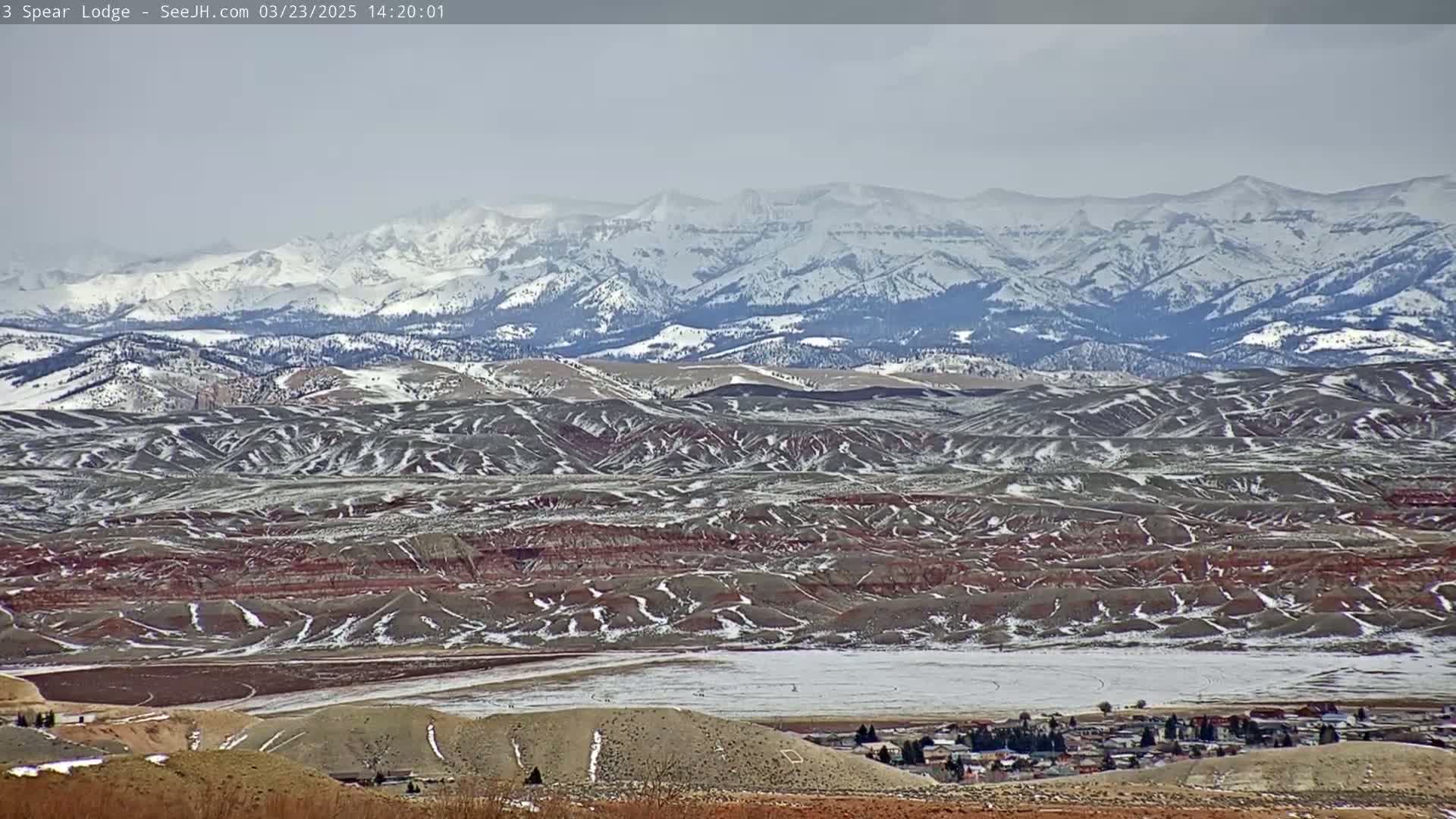 A snow-dusted landscape featuring rolling hills and a small town in the foreground, with a distant mountain range covered in snow under an overcast sky.
