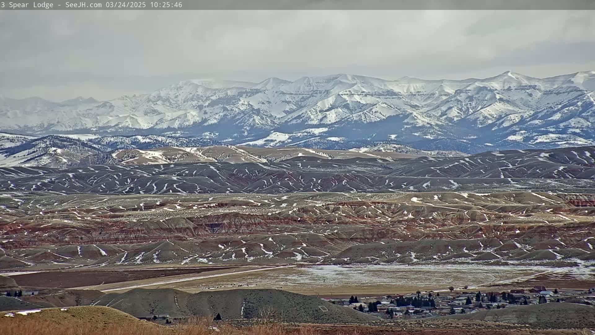 A snow-dusted valley with rolling hills and a small town in the foreground, overlooks a distant mountain range capped with snow under an overcast sky.
