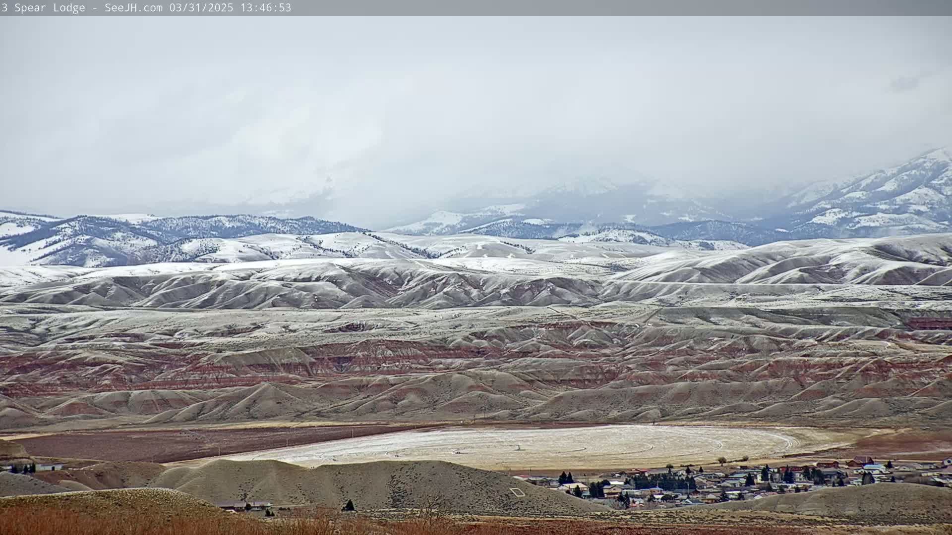 A snow-dusted landscape of rolling hills and distant mountains under an overcast sky stretches out to a small town in a valley.
