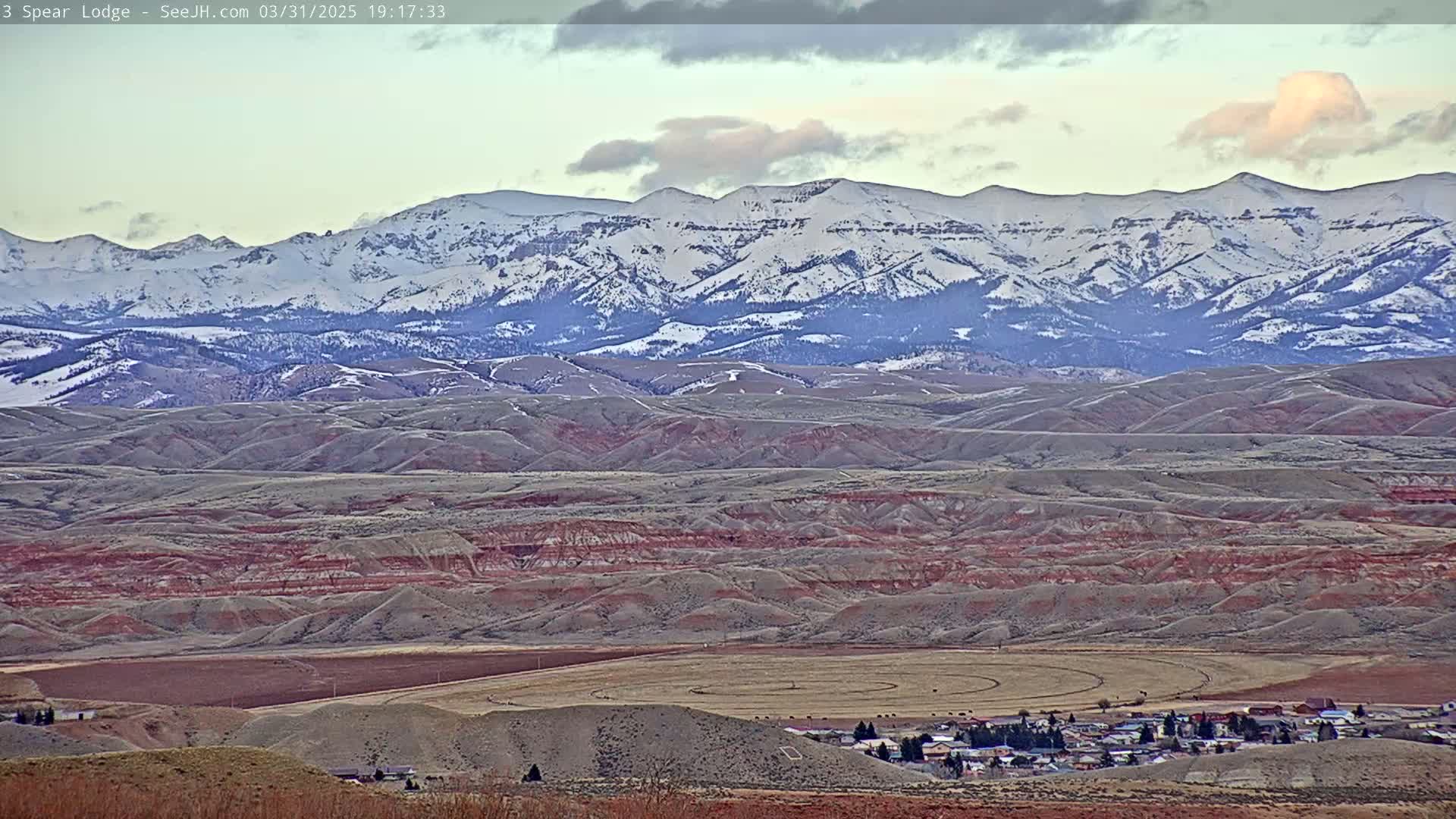 A small town nestled in a valley of rolling, reddish-brown hills, with a snow-capped mountain range in the background under a partly cloudy sky.