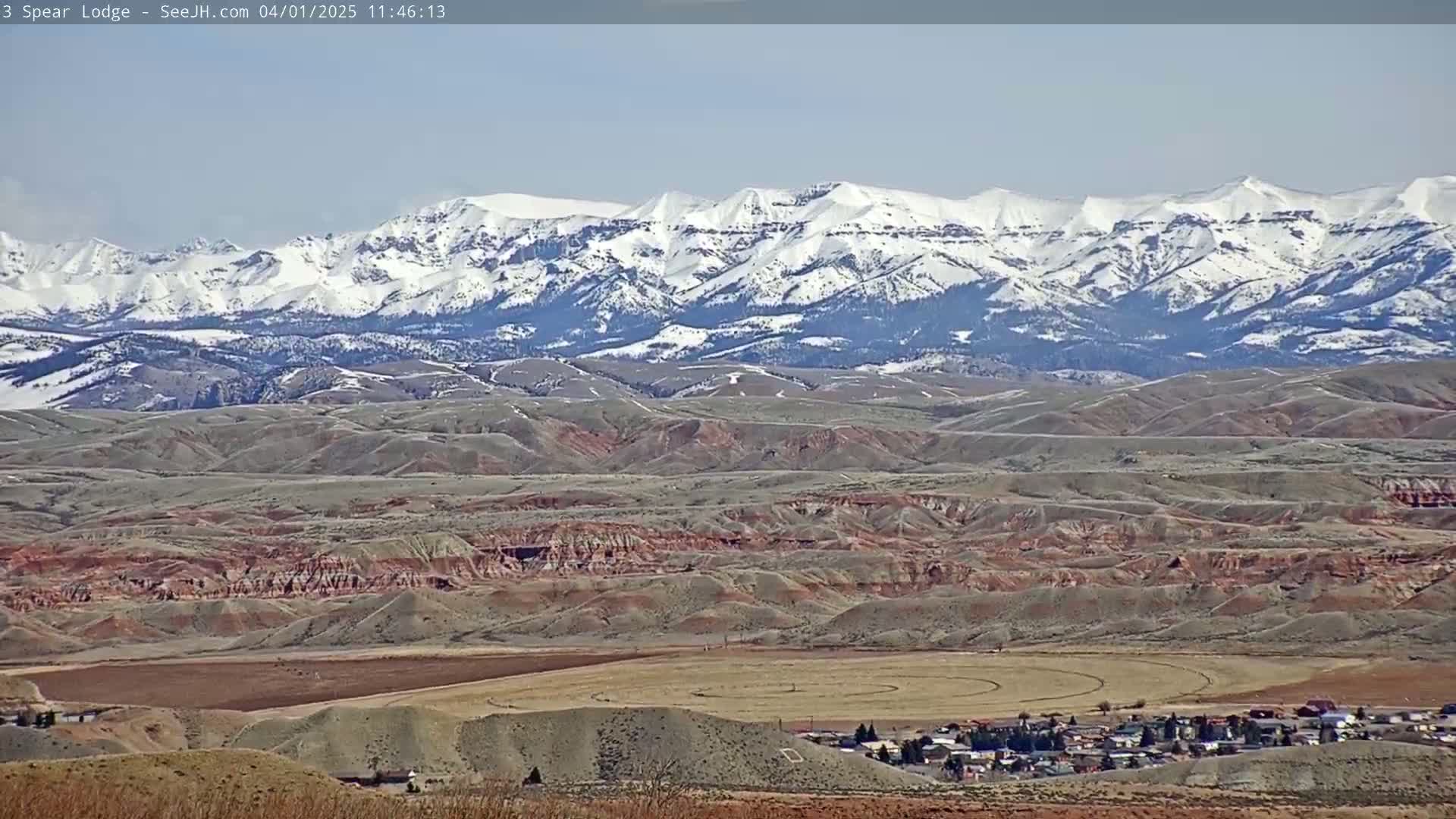 A long-range view shows a snow-capped mountain range in the background, a valley with rolling hills and reddish-brown earth in the mid-ground, and a small town nestled in the foreground under a clear sky.