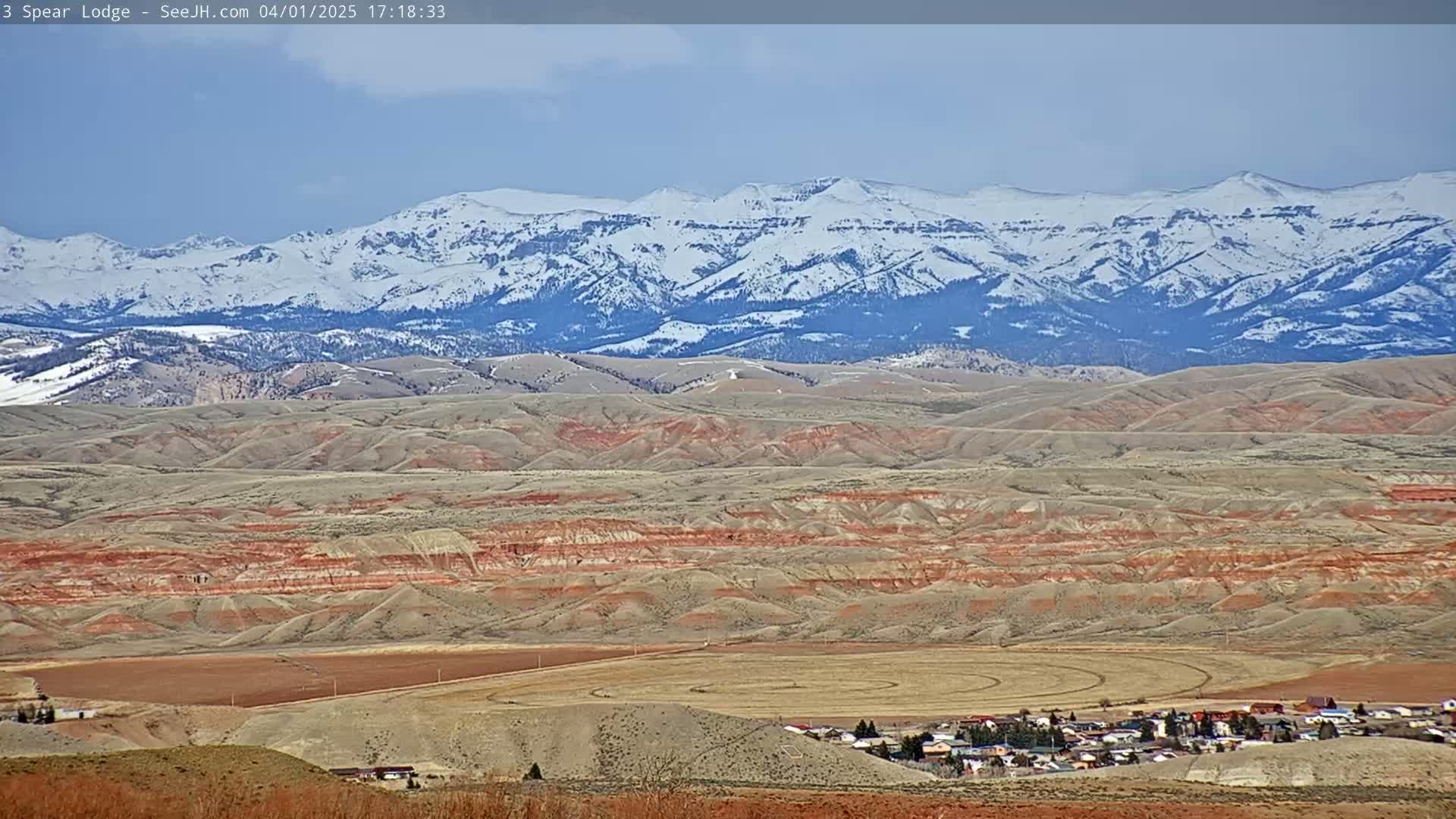 A snow-capped mountain range is visible in the background of a landscape featuring rolling hills of reddish-brown earth and a small town nestled in a valley.