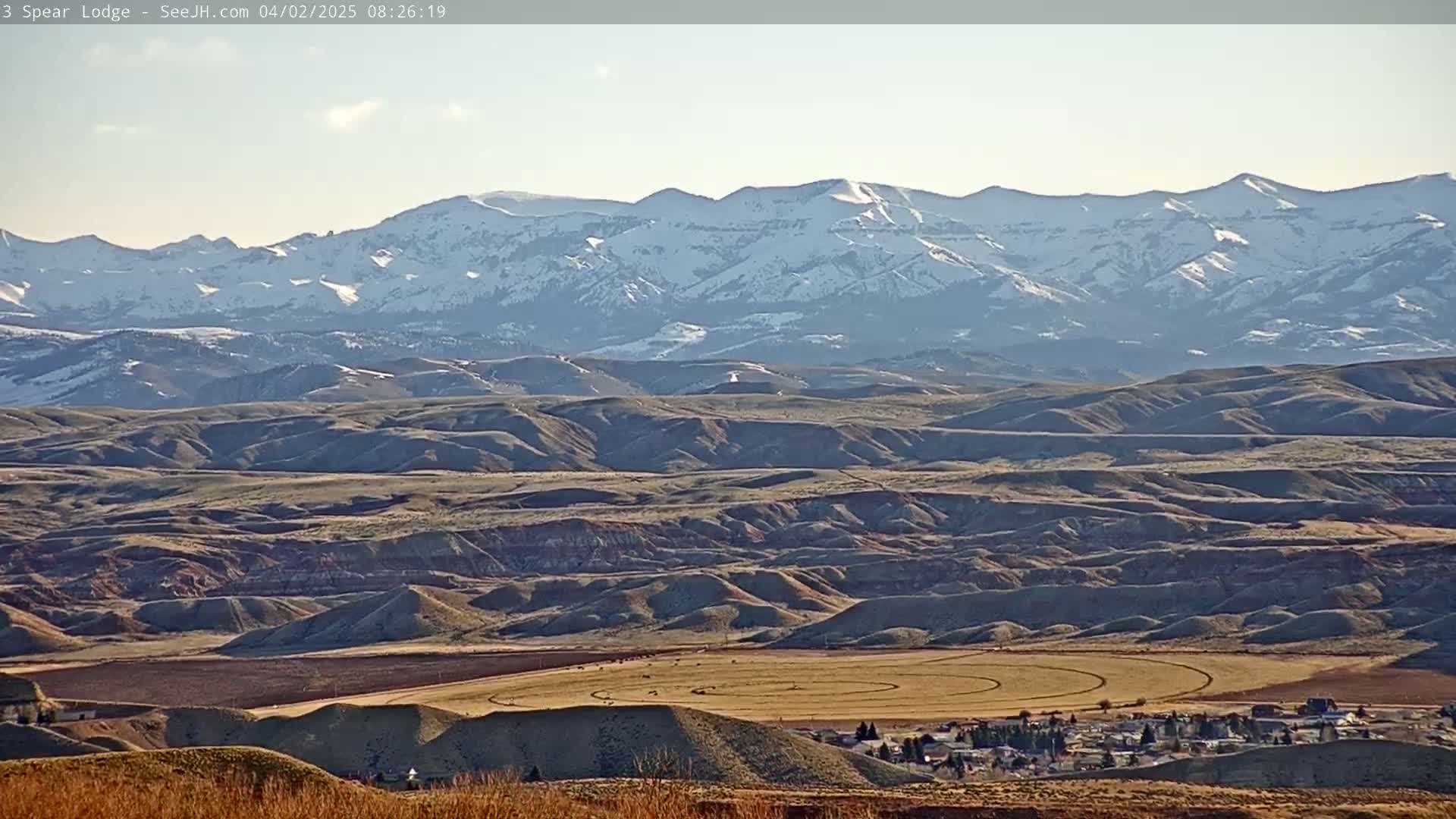 A vast, undulating landscape of brown and tan hills stretches towards a snow-capped mountain range under a clear, bright sky.