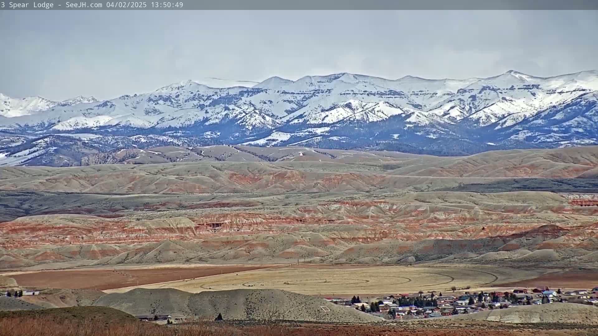 A small town nestled in a valley of rolling, reddish-brown hills, with a snow-capped mountain range in the background under a mostly cloudy sky.