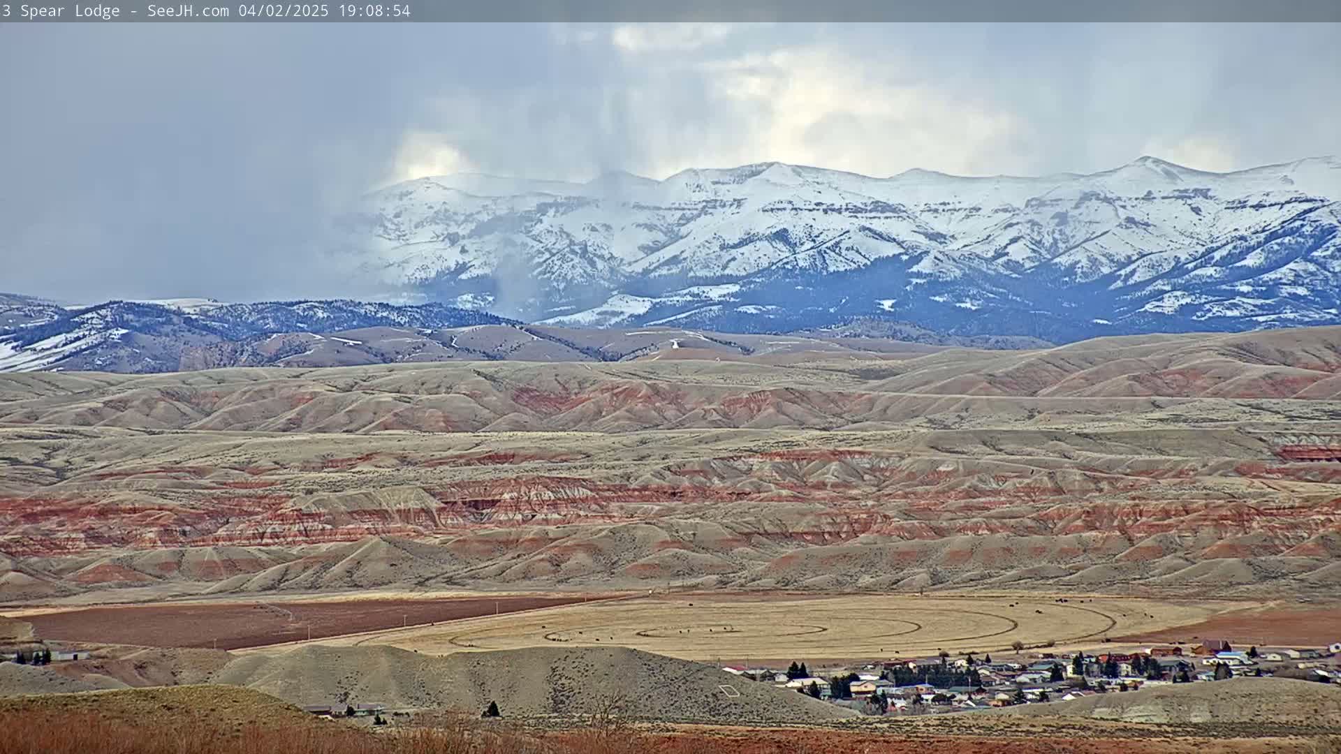 A snow-capped mountain range is visible in the background of a vast, undulating landscape of tan and reddish-brown hills, with a small town and a circular field in the foreground under a cloudy sky.