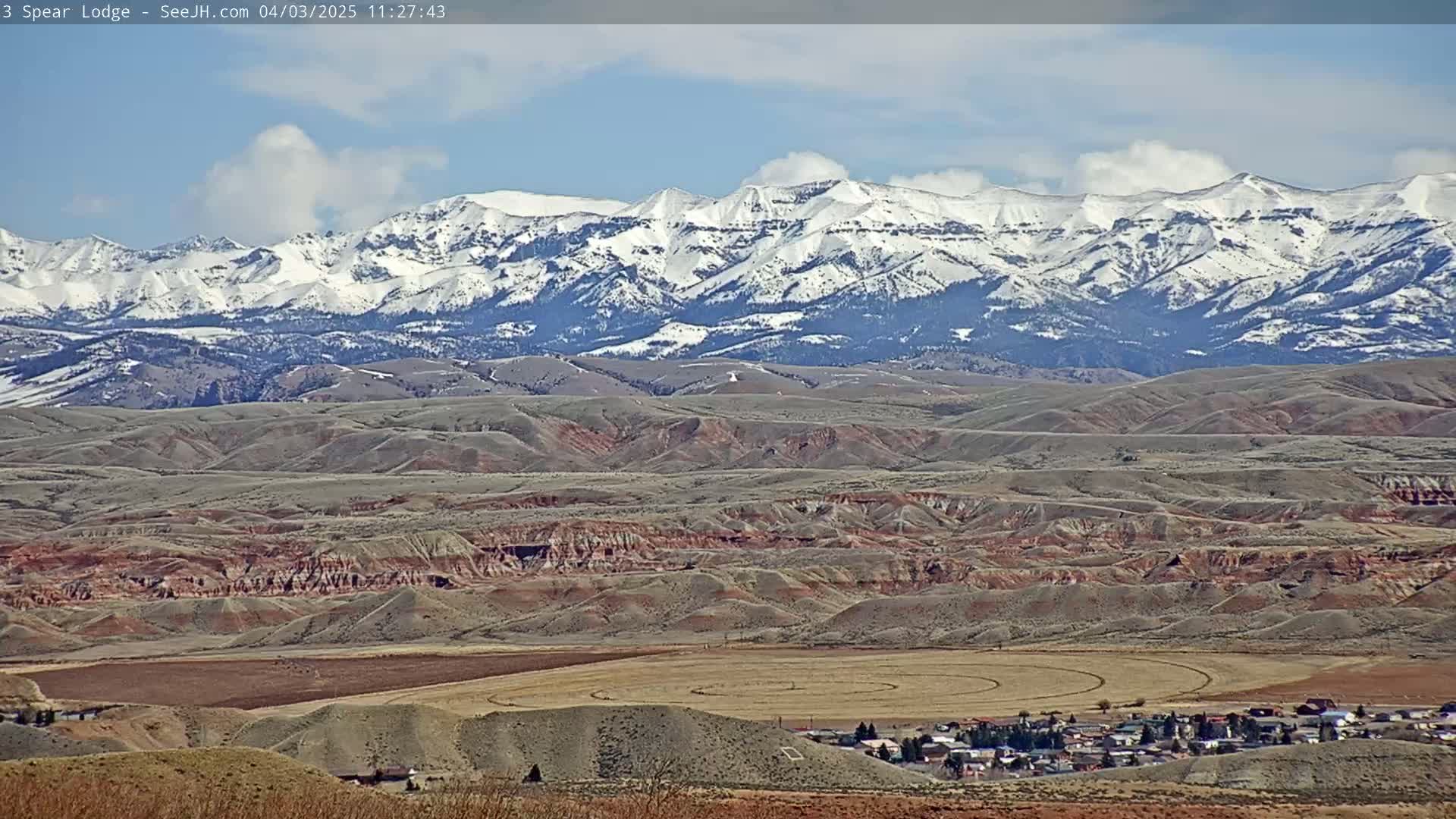 A small town sits in a valley of reddish-brown hills, with a snow-capped mountain range in the background under a partly cloudy sky.