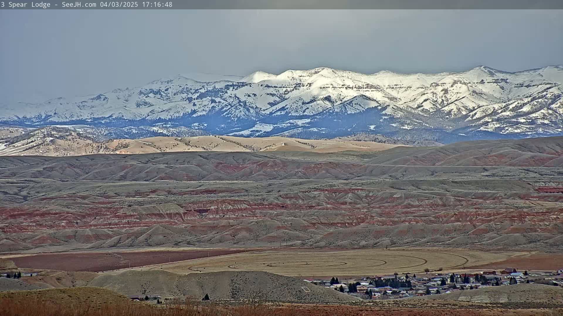 A small town nestled in a valley of rolling, reddish-brown hills, with a snow-capped mountain range in the background under an overcast sky.
