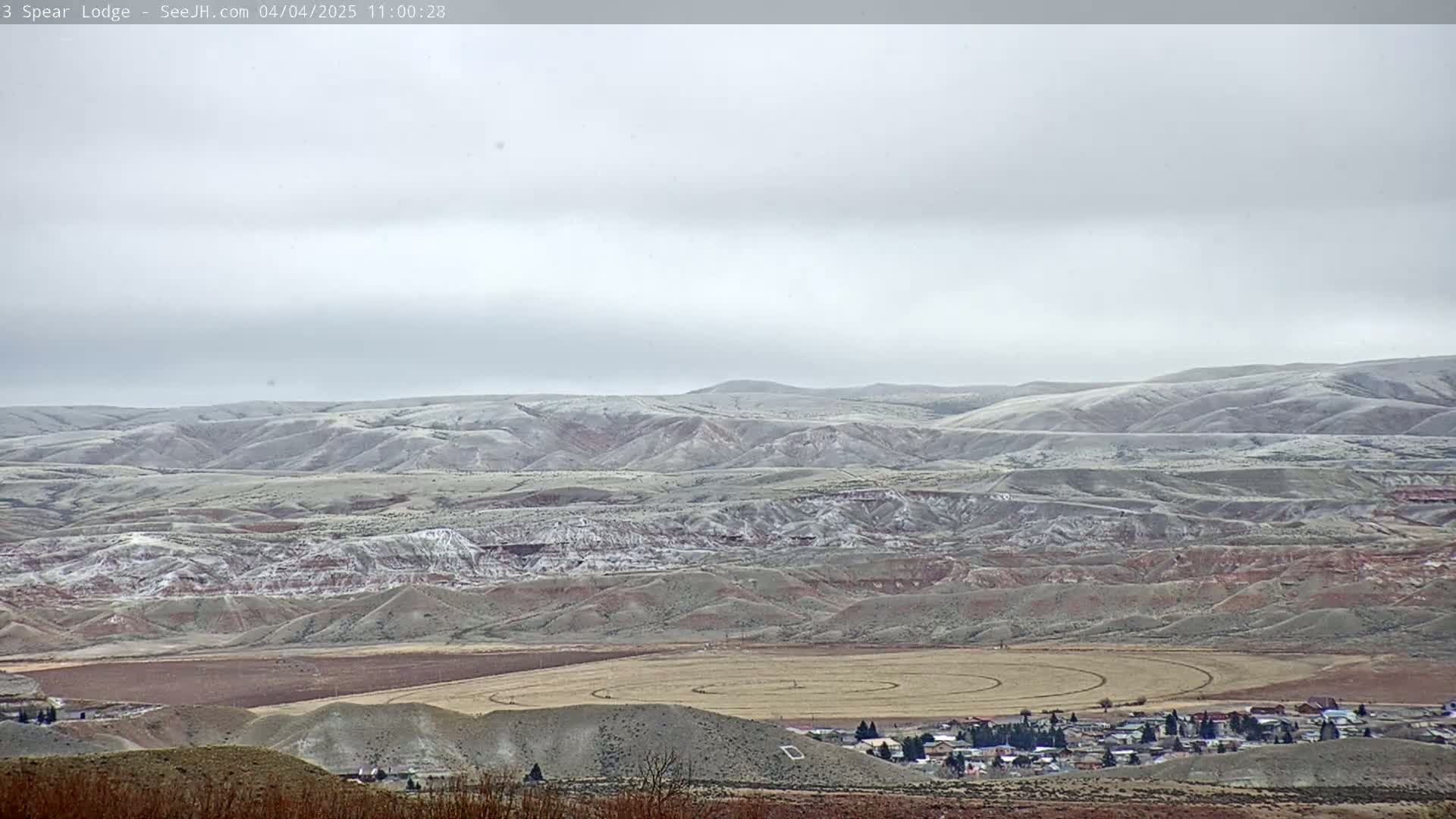 A snow-dusted, hilly landscape with a small town and a circular field in the foreground under an overcast sky.