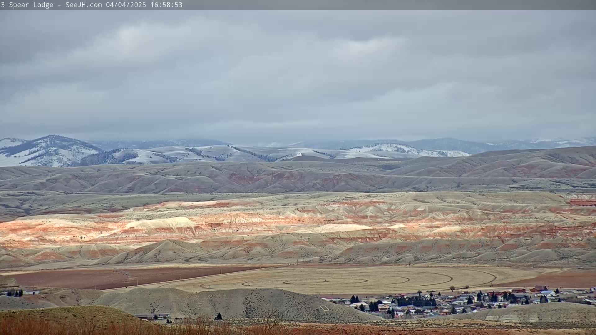 A long-range view shows a small town nestled in a valley of rolling hills and distant mountains with patches of snow under a cloudy sky.