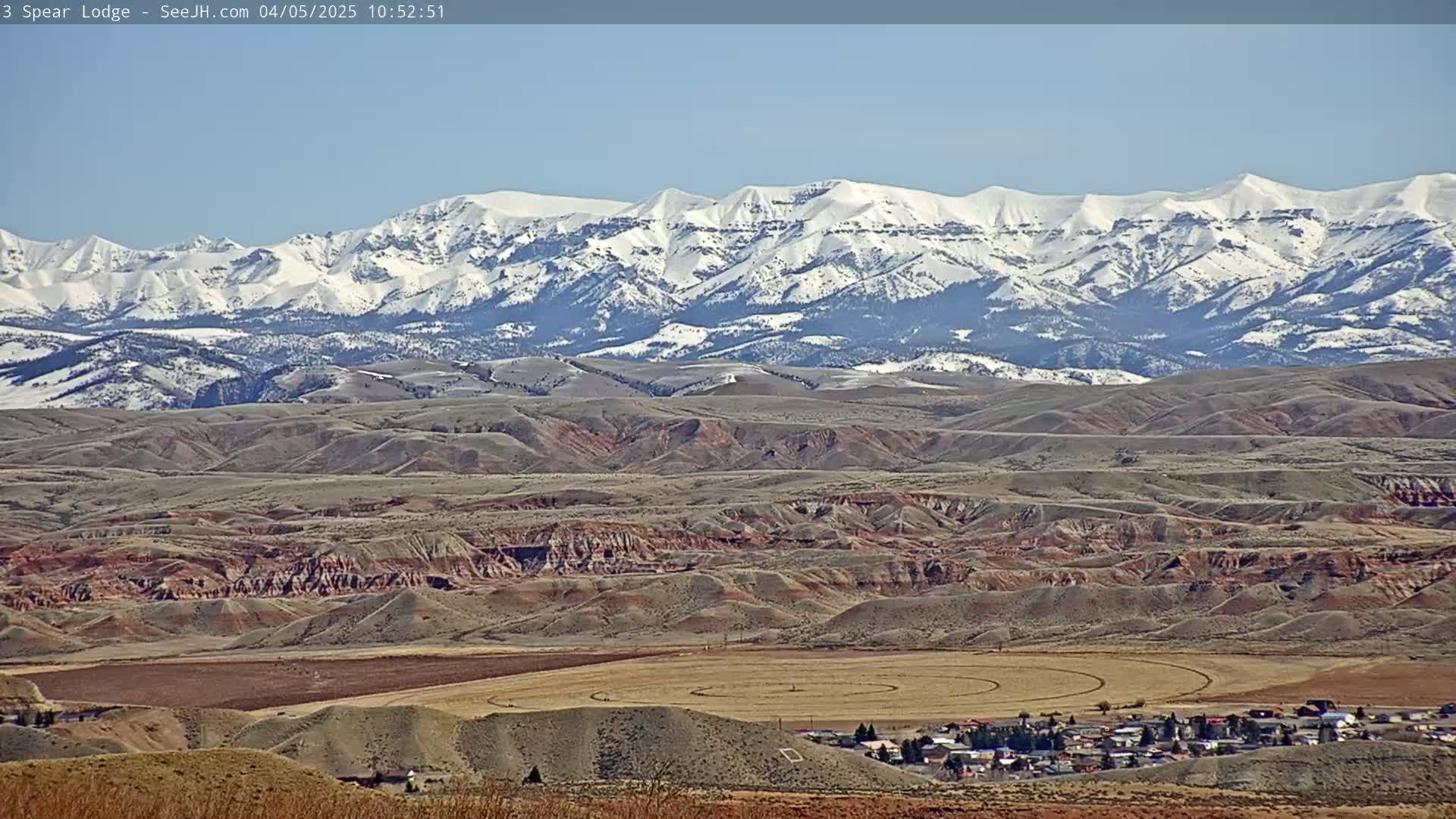A long-range view shows a snow-capped mountain range in the background, overlooking a vast expanse of rolling hills and a small town, under a clear, sunny sky.