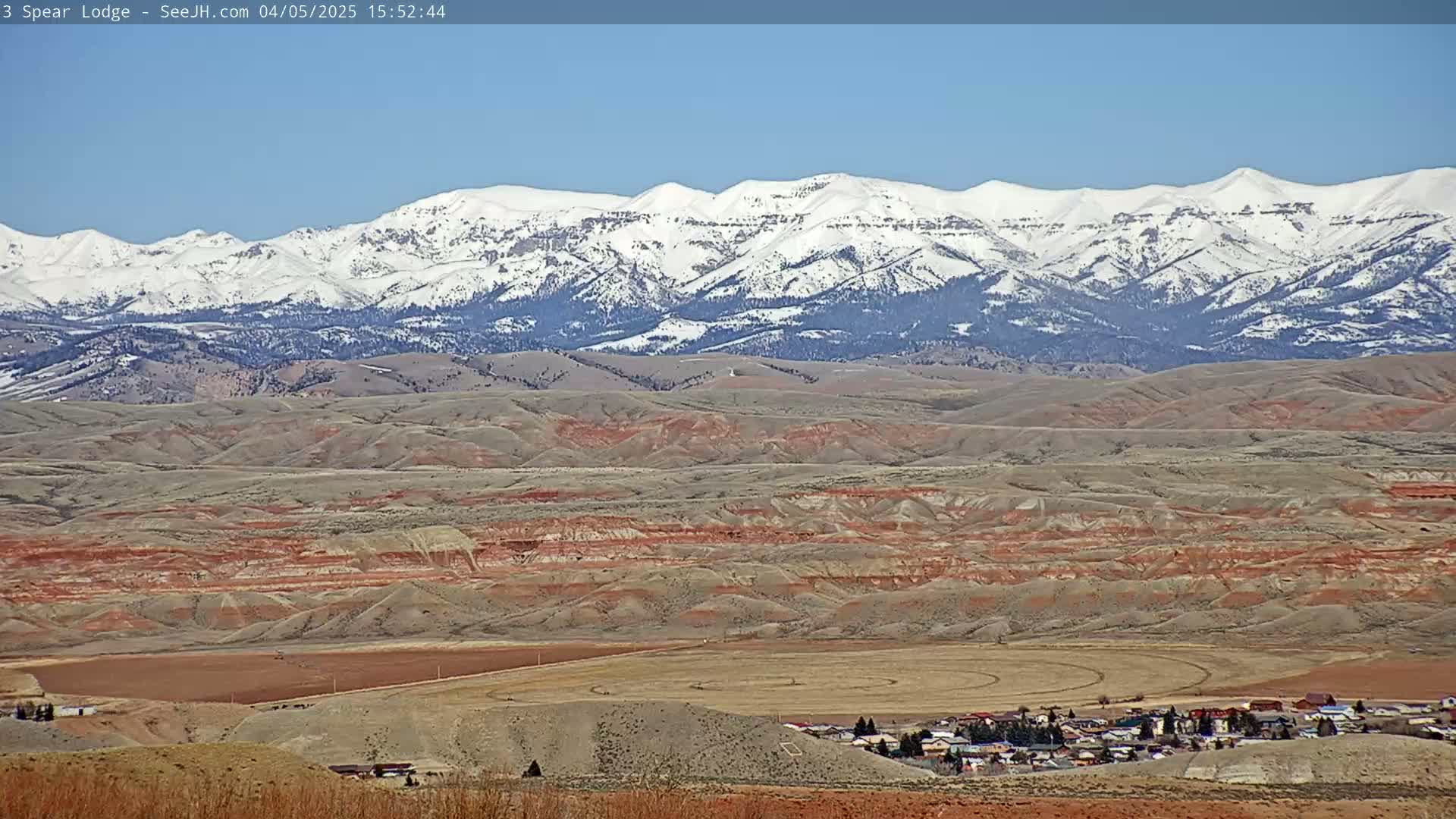 A small town sits in a valley of reddish-brown hills, with a snow-capped mountain range in the background under a clear blue sky.