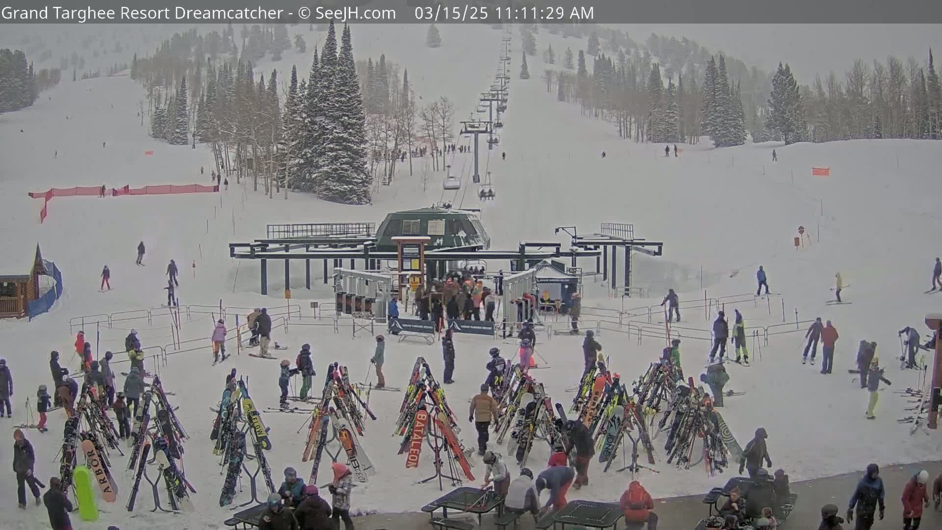 A ski resort base area is covered in snow during a snowfall, with skiers and snowboarders gathered around racks of skis and snowboards near a chairlift.