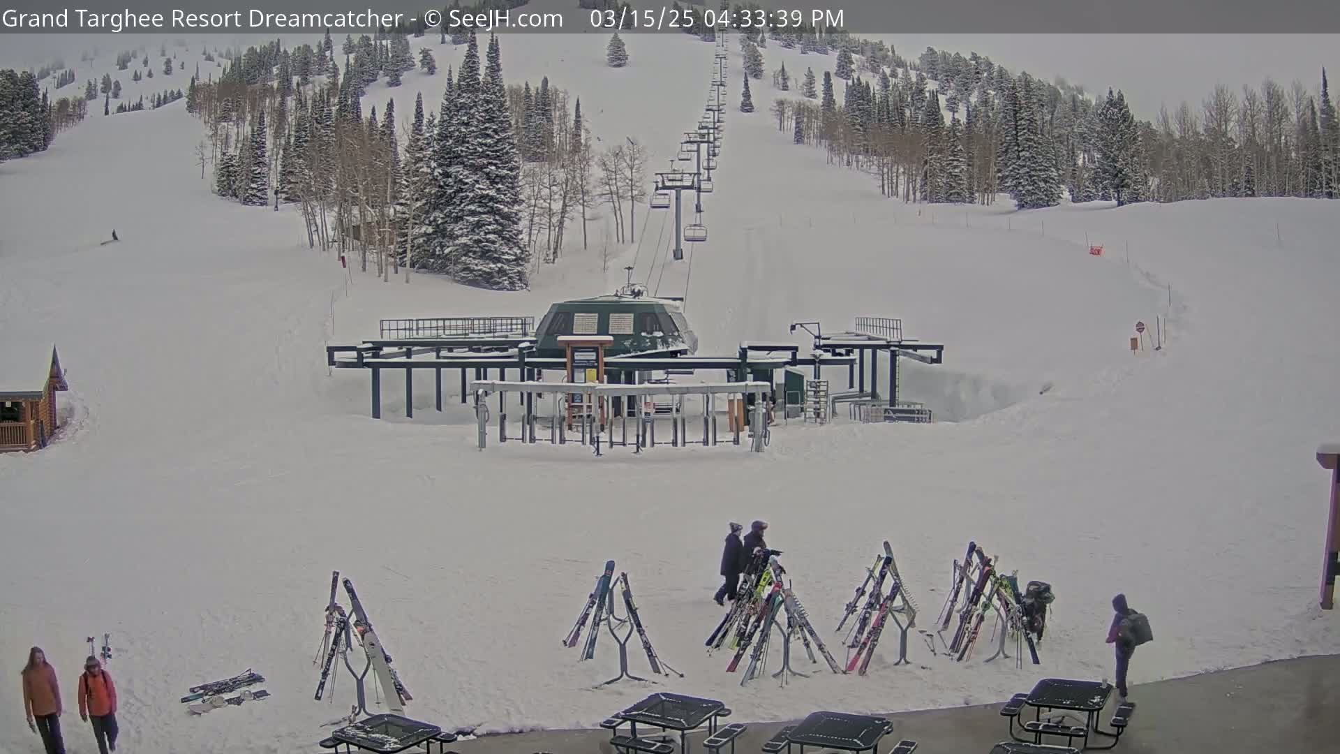 A ski lift base area covered in snow, with several pairs of skis leaning against racks near picnic tables, and a few people walking around.