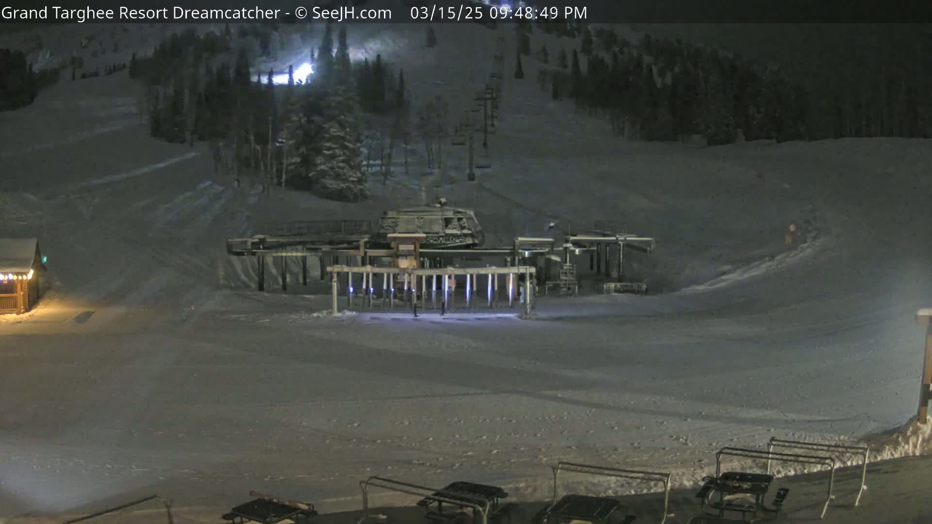 A nighttime view of a snow-covered ski slope with a chairlift and several picnic tables at the bottom.