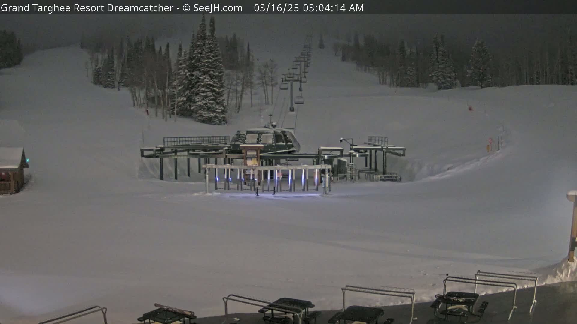 A ski lift at night sits in a snow-covered area with a line of chairs extending uphill, under a dark sky.