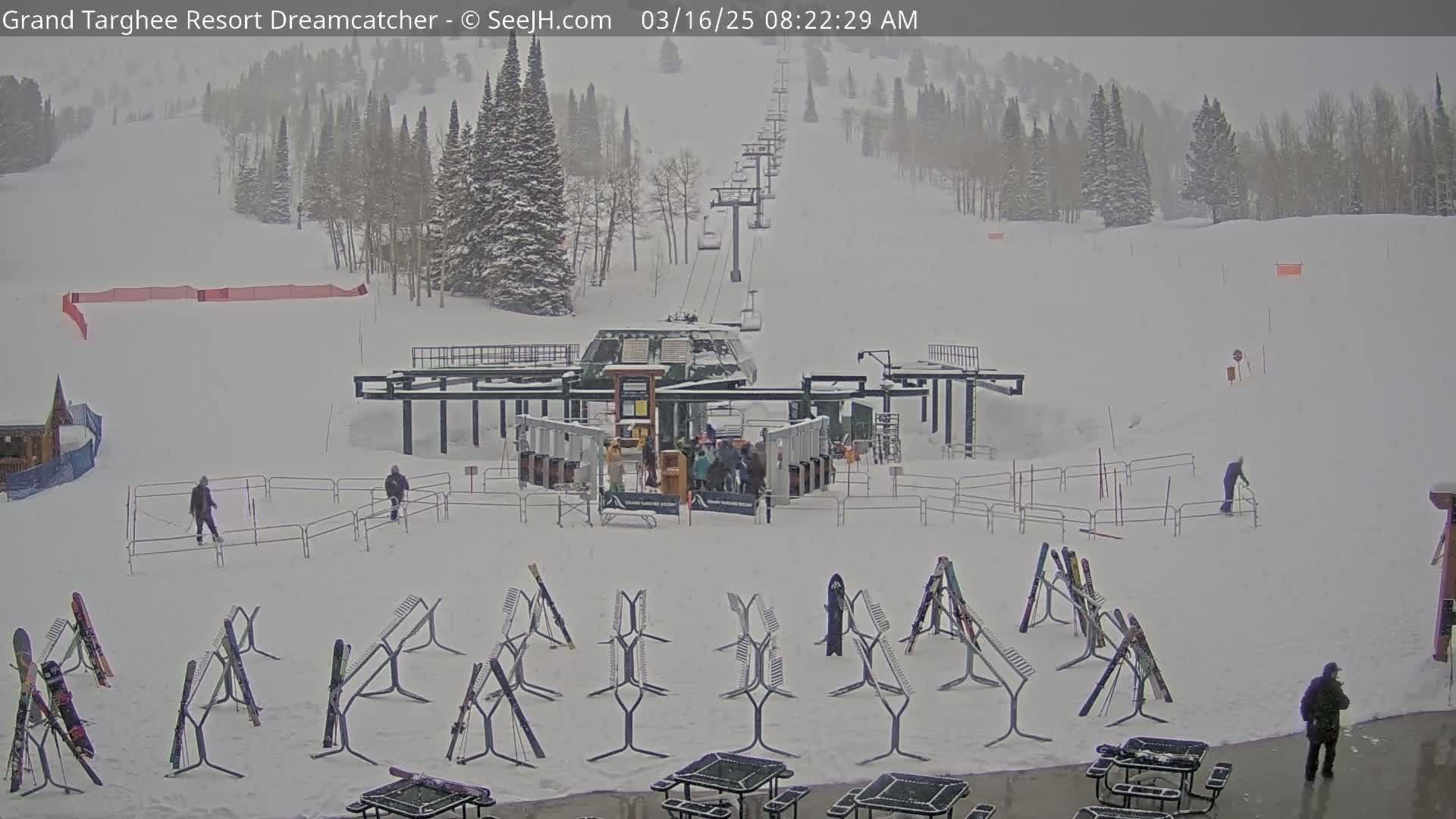 A ski lift base area is covered in snow during a snowfall, with skiers and snowboarders present, and several ski racks holding various equipment.