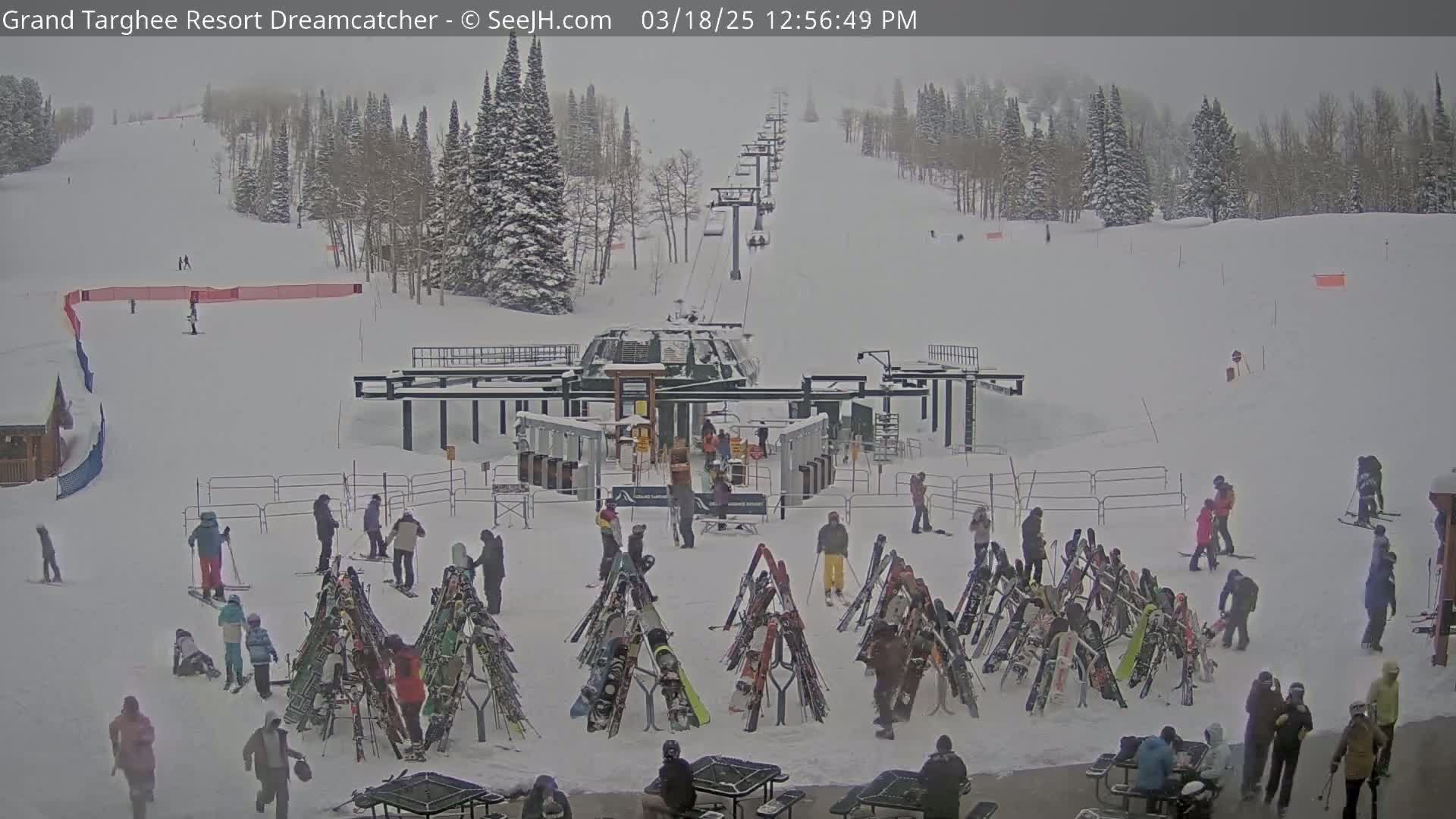 A snowy ski resort base area shows a chairlift, skiers and snowboarders, and numerous skis and snowboards leaning against racks.