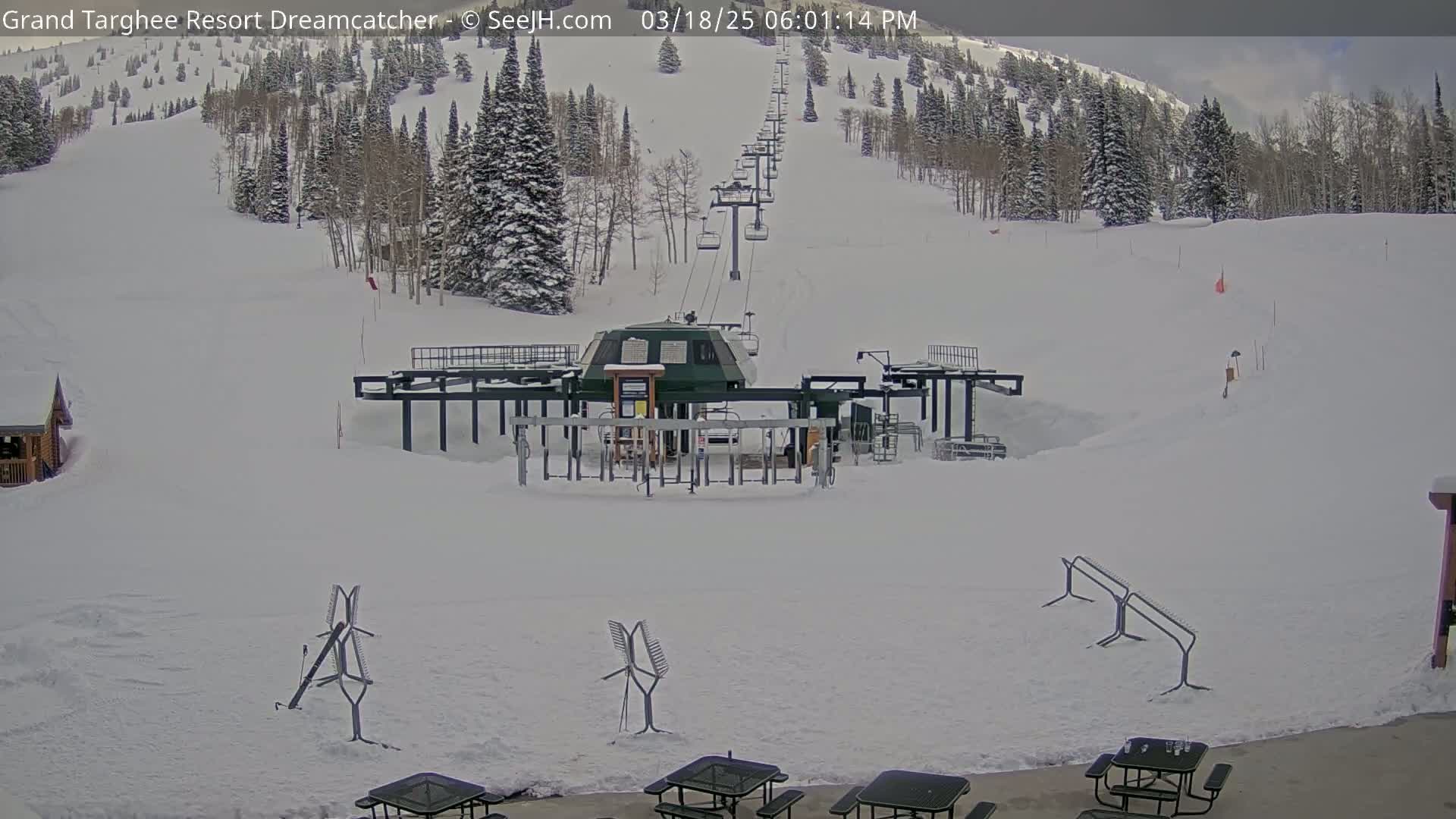 A snow-covered ski lift base area is seen with a chairlift ascending a snow-covered mountainside under a partly cloudy sky.