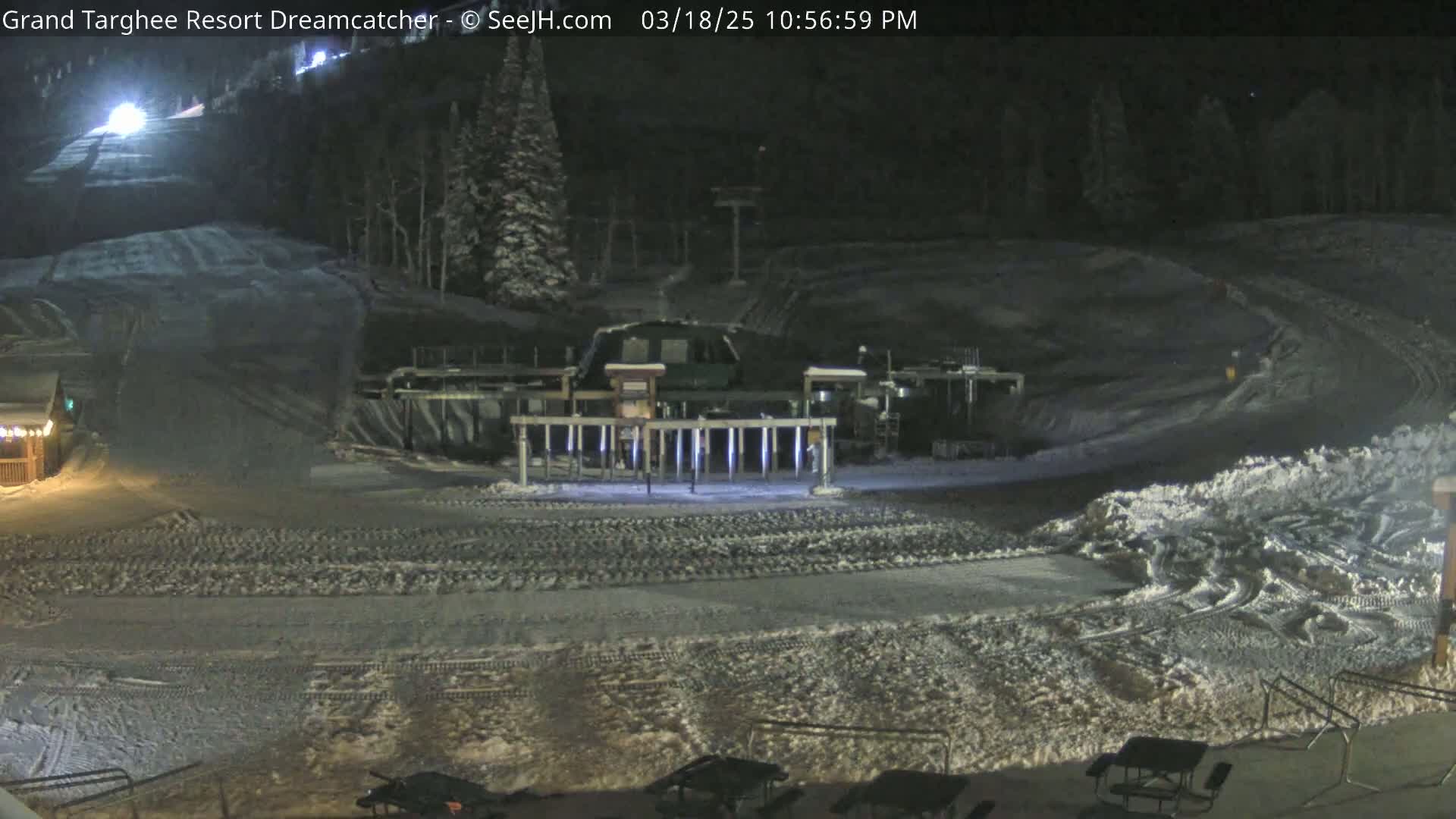 A snow-covered ski resort at night, illuminated by artificial lights, showing a lift and a building, with tire tracks visible on the slopes.