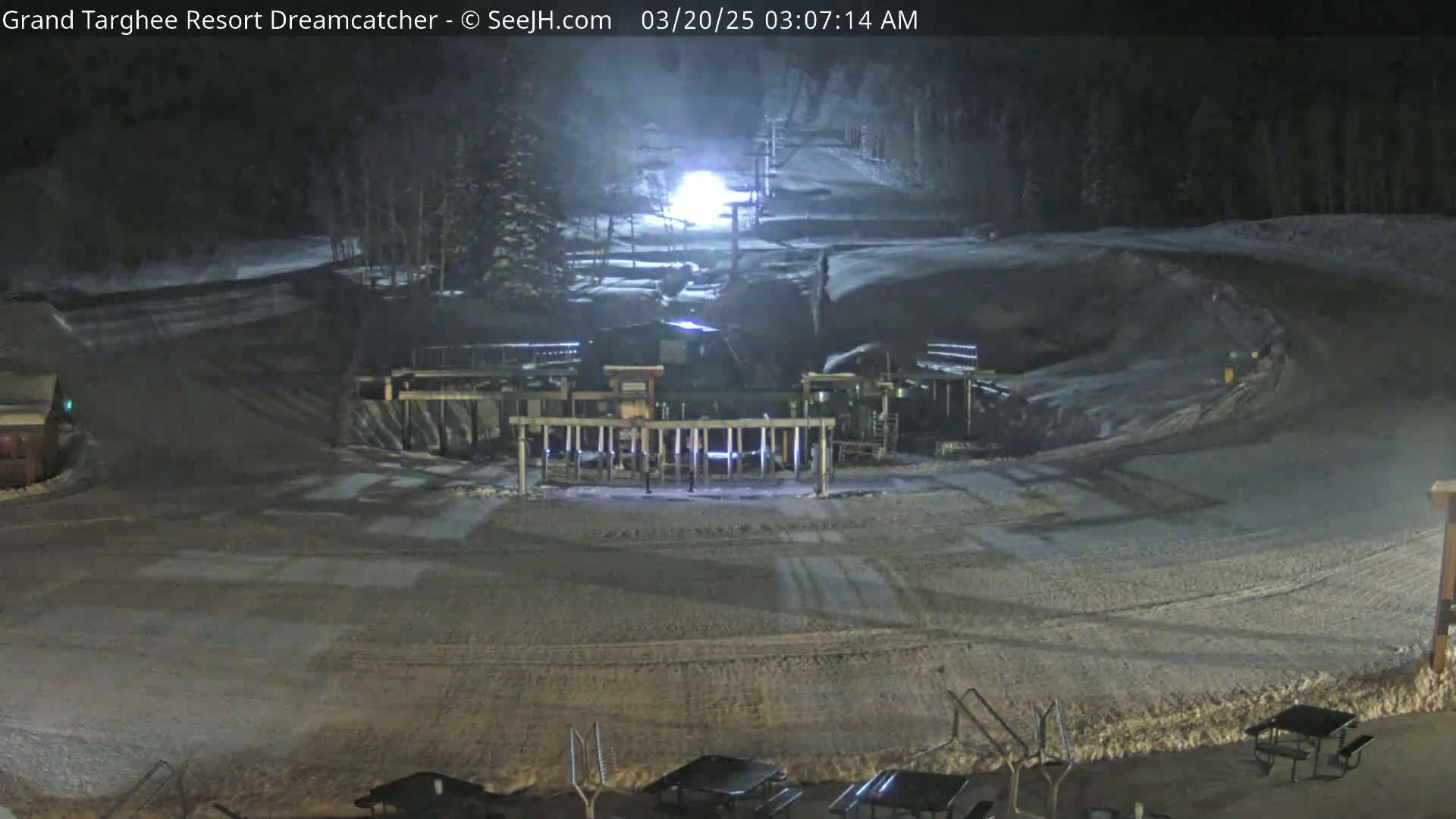 A snow-covered ski resort at night, illuminated by artificial lights, shows a ski lift and several picnic tables.