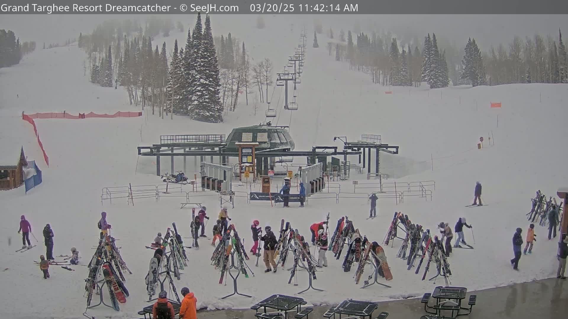 A ski lift base area with numerous skiers and snowboarders around ski racks in snowy conditions during a light snowfall.