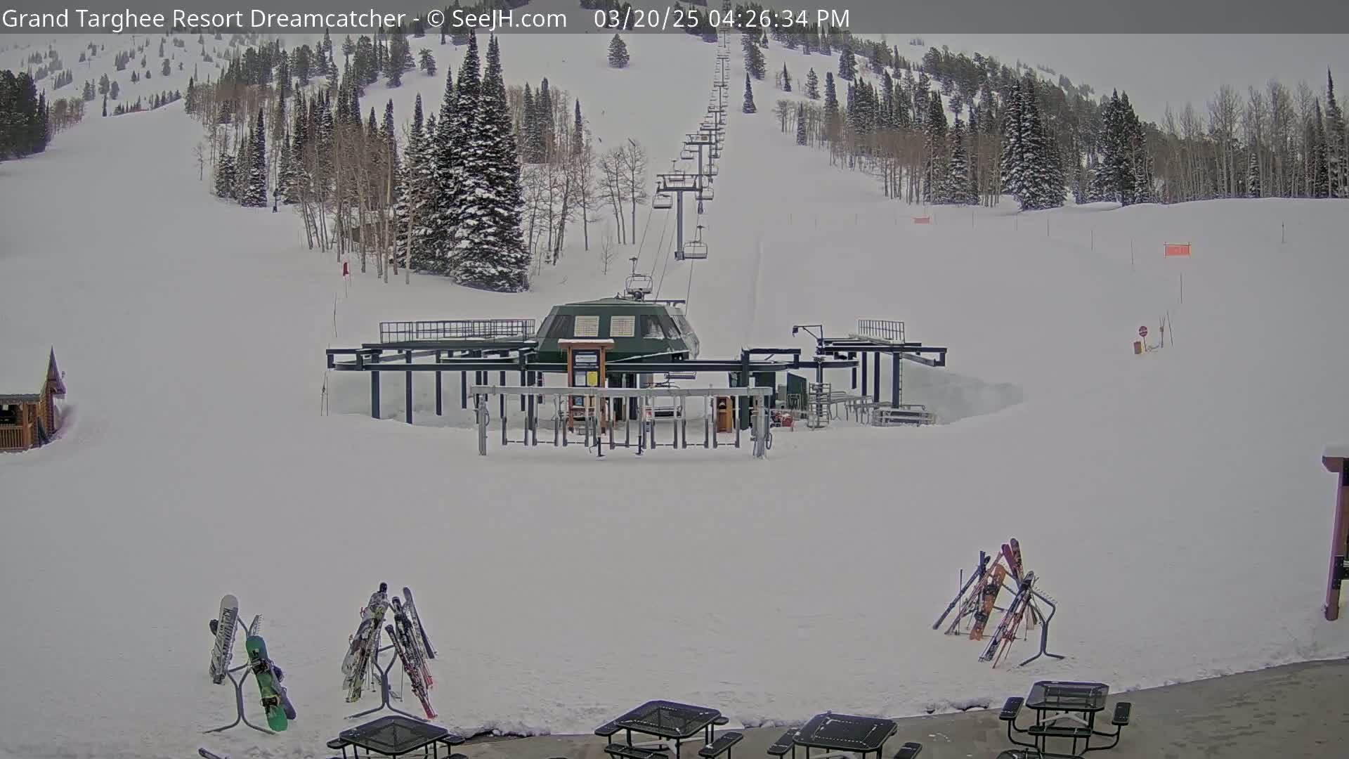 A ski lift base station sits in a snowy landscape with a ski run extending uphill to a wooded area under an overcast sky.