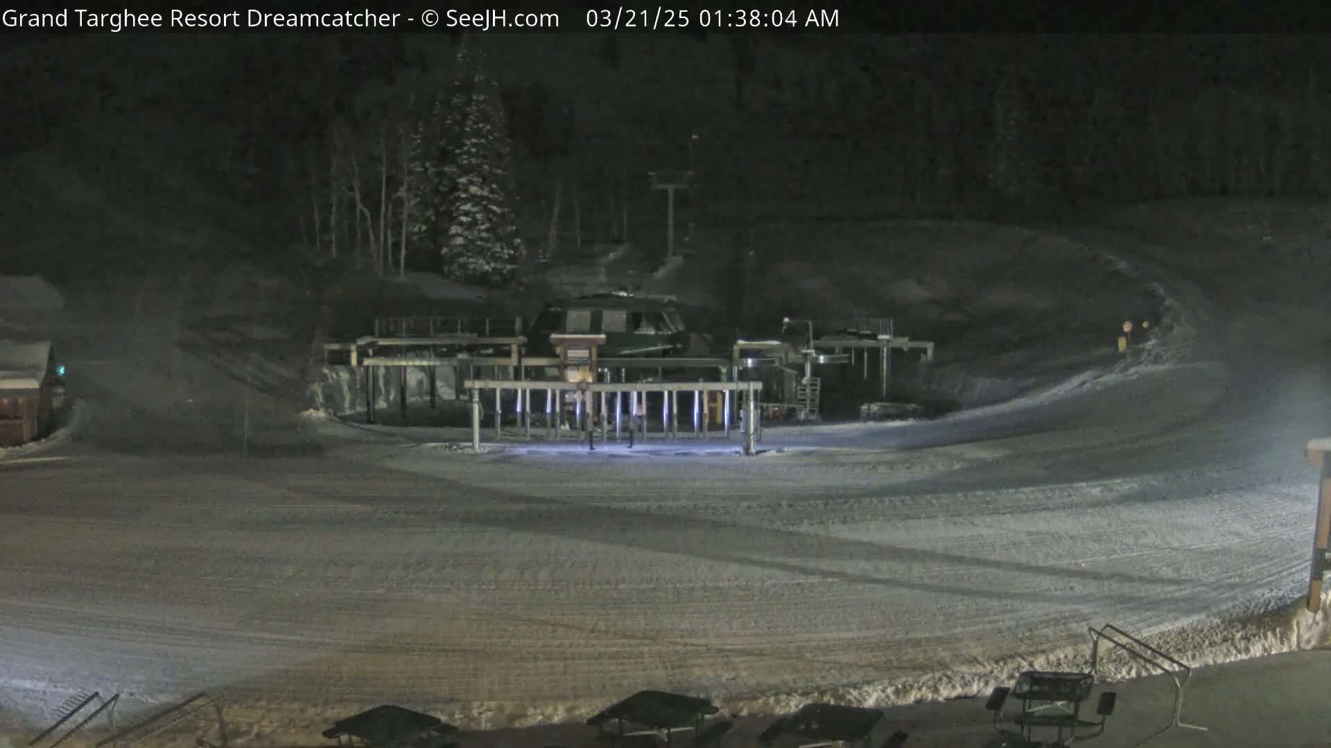 A snow-covered ski resort at night, illuminated by artificial lights, shows a ski lift and a few people near it.