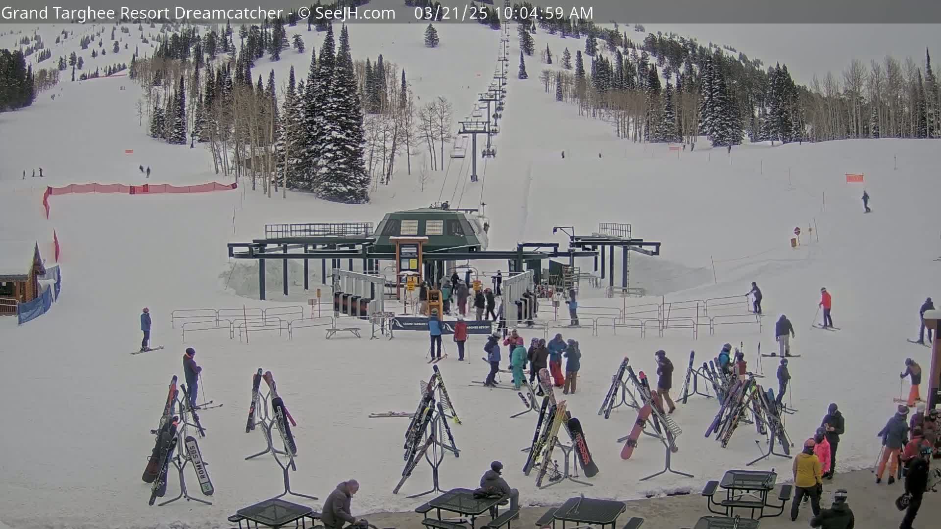A ski lift base area with numerous skiers and snowboarders, many skis and snowboards leaning against racks, and several people congregating, all under an overcast sky.