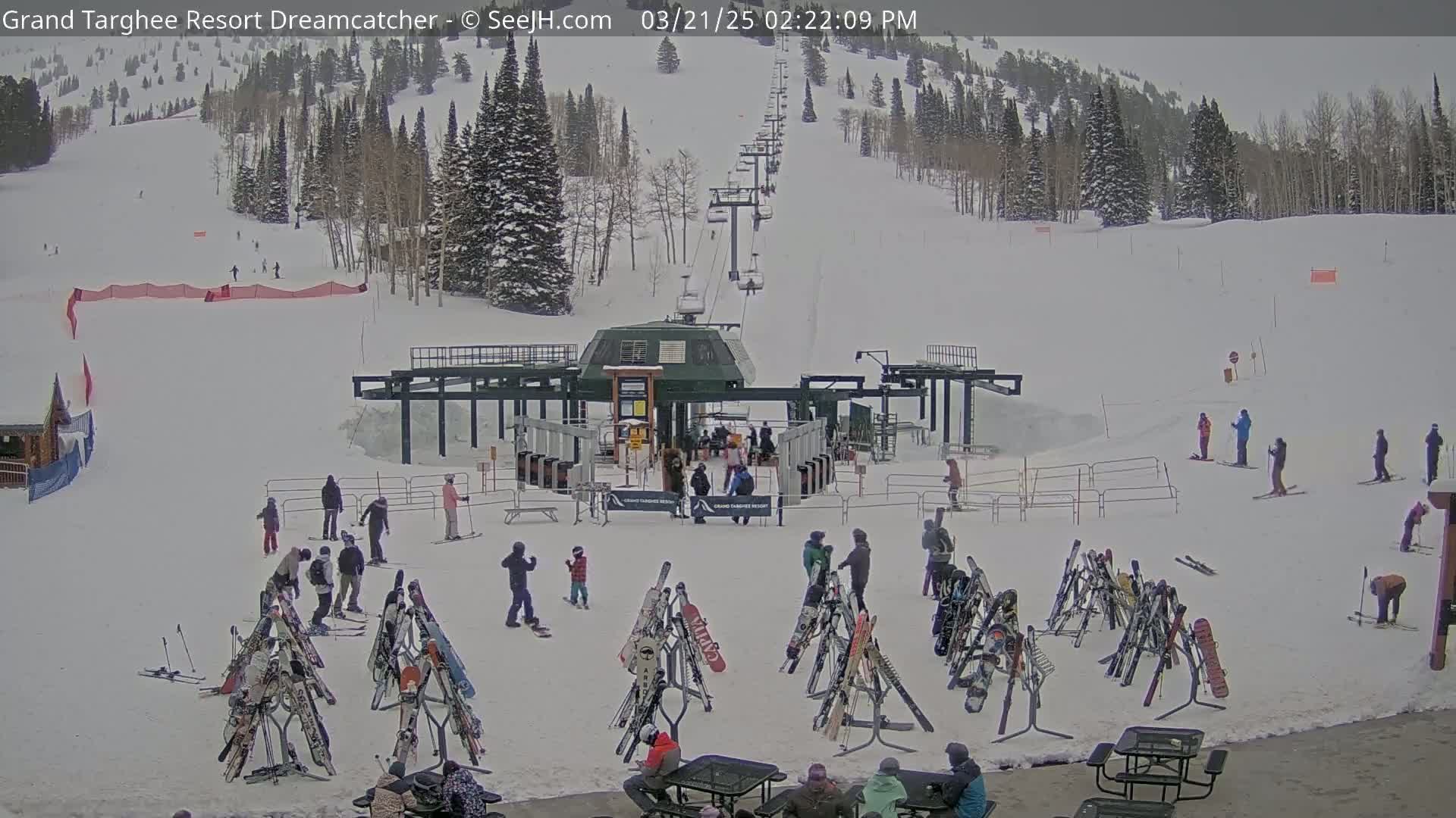 A ski resort base area shows a ski lift, numerous skiers and snowboarders, and several ski racks on a snowy day.