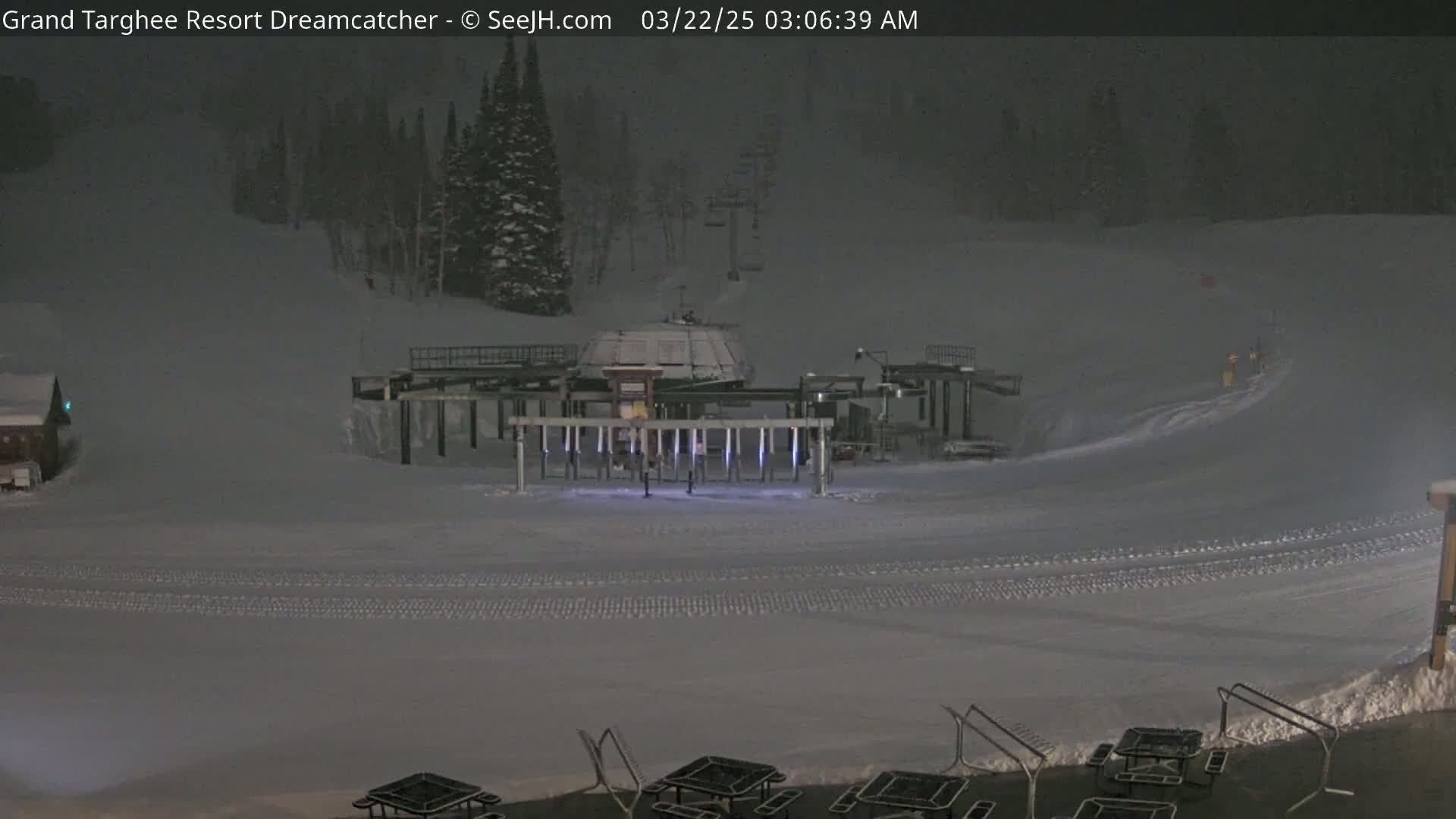 A snow-covered ski resort at night, lightly snowing, shows a ski lift and several empty picnic tables.