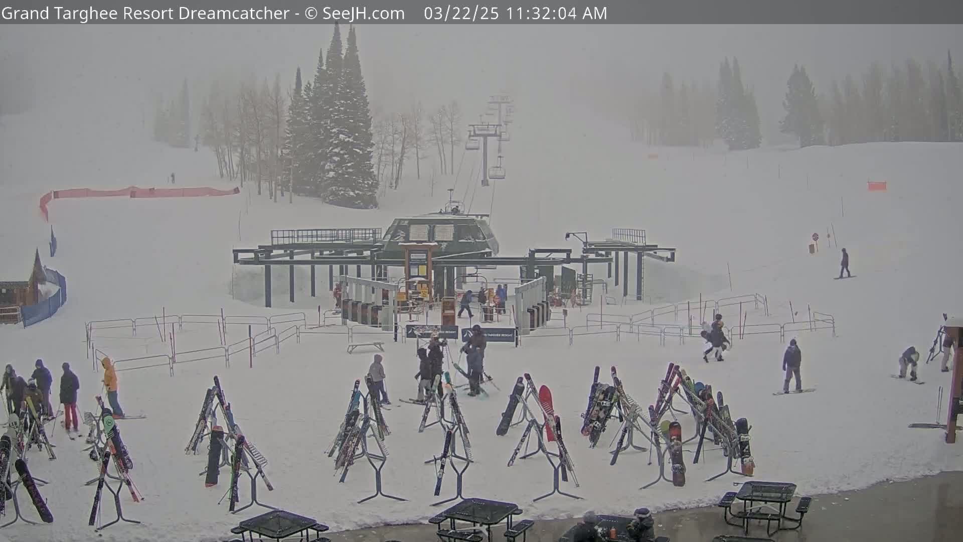 A snowy ski resort base area with a chairlift, skiers and snowboarders, and numerous skis and snowboards resting on racks in foggy conditions.