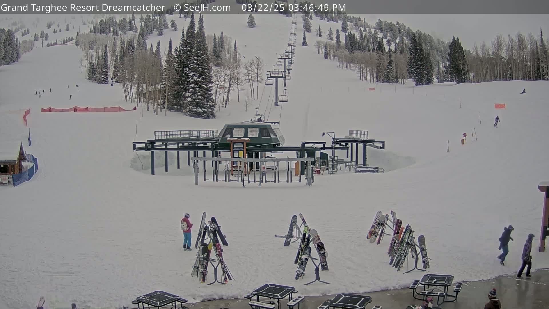 A ski lift base area with snow-covered slopes and several people, some carrying skis, under an overcast sky.